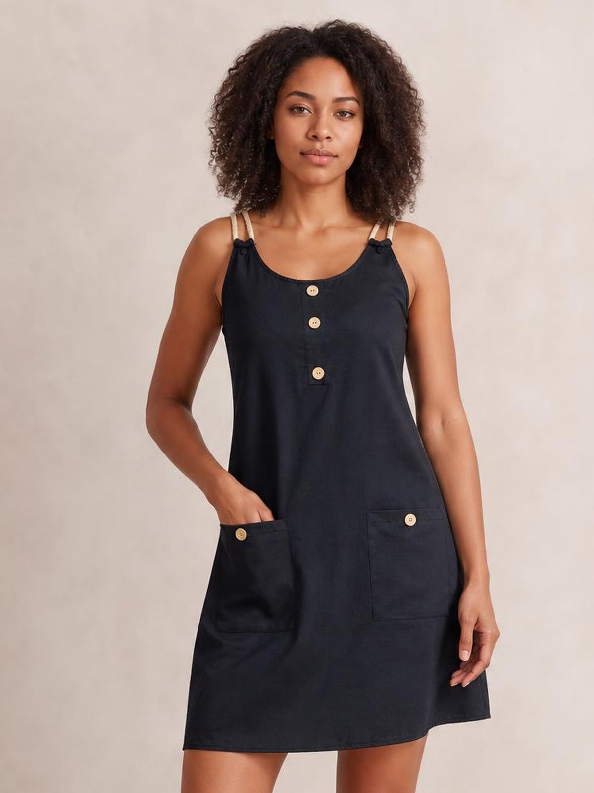 Model wearing a black round-neck mini dress with double spaghetti straps, decorative front buttons, two patch pockets, and a relaxed A-line silhouette.