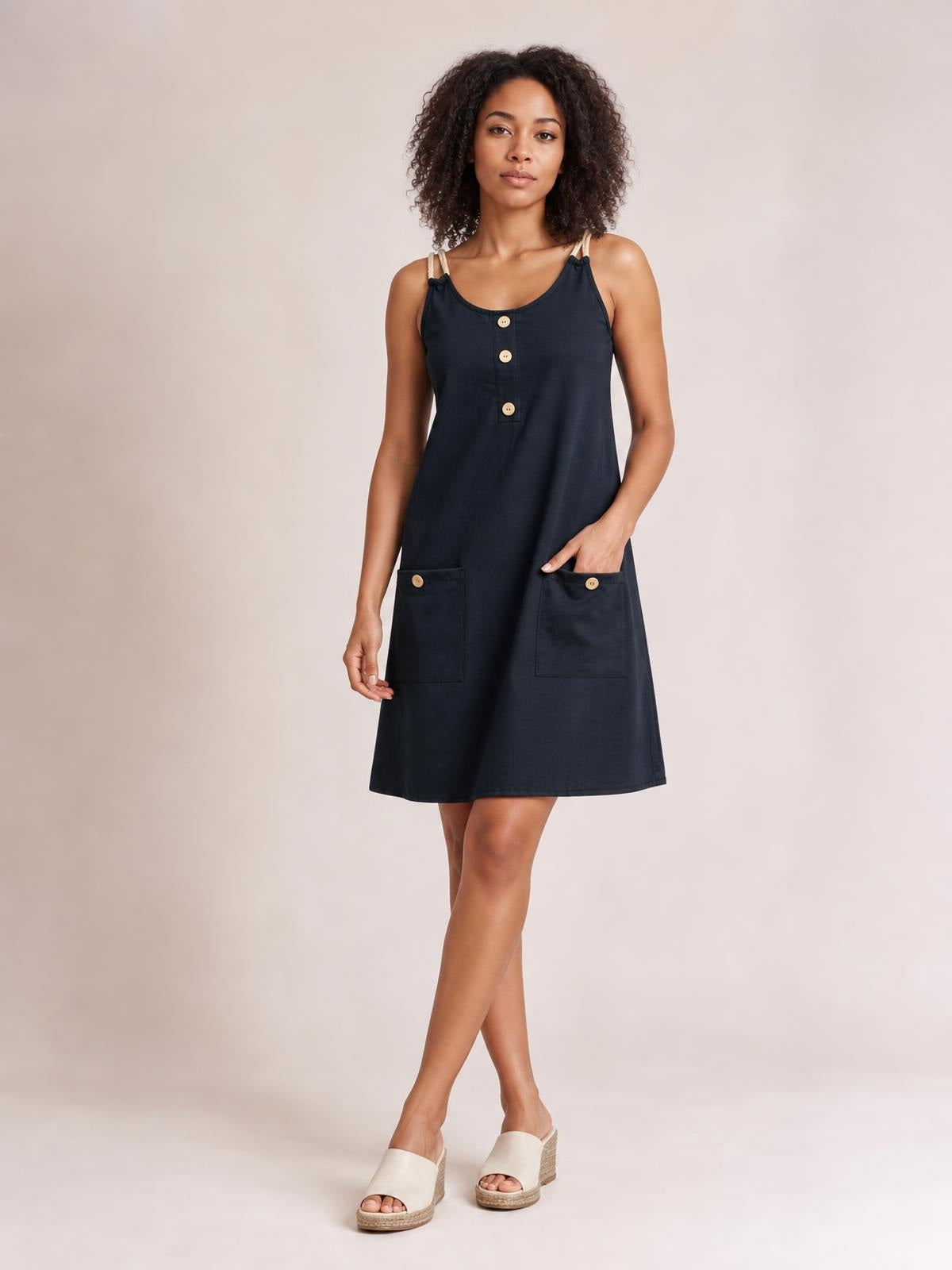Model wears a navy round-neck mini dress with double spaghetti straps, decorative front buttons, patch pockets, and a relaxed A-line silhouette while standing cross-legged.
