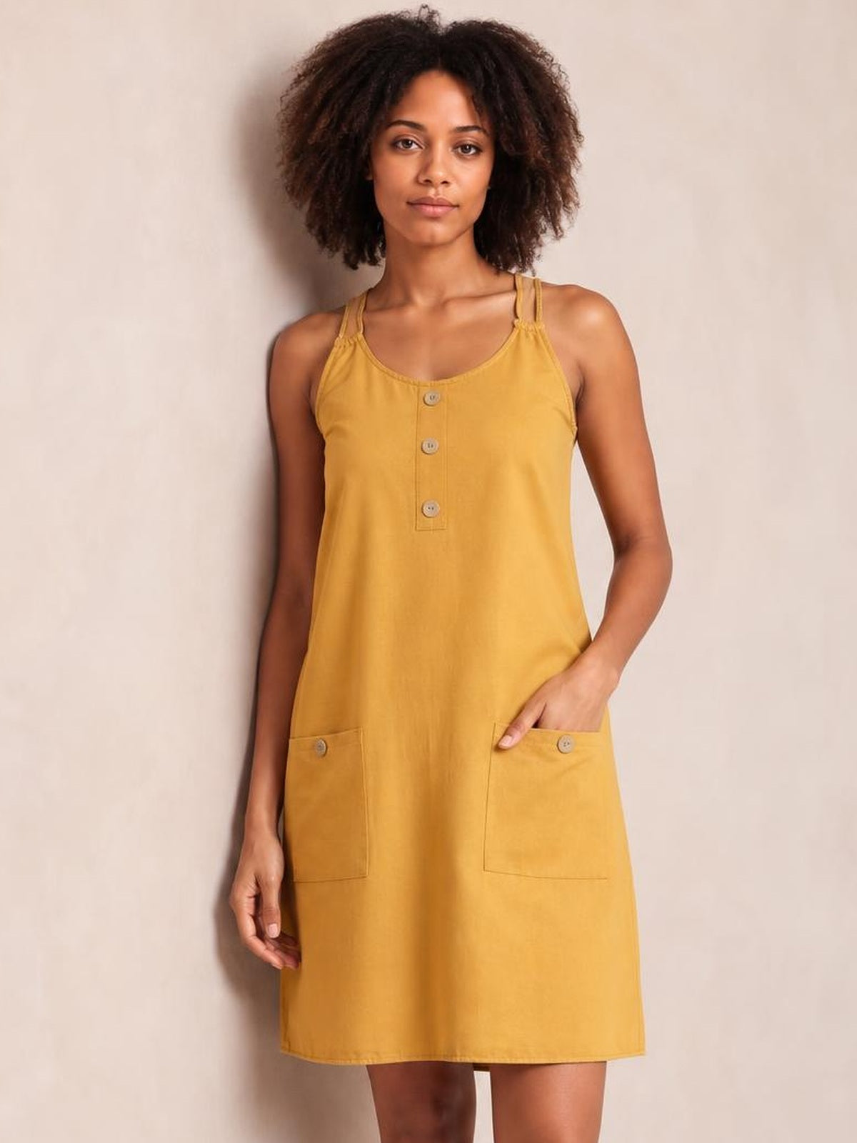 A model stands in a mustard-yellow round-neck mini dress with double spaghetti straps, a front button placket, straight silhouette, and patch pockets.