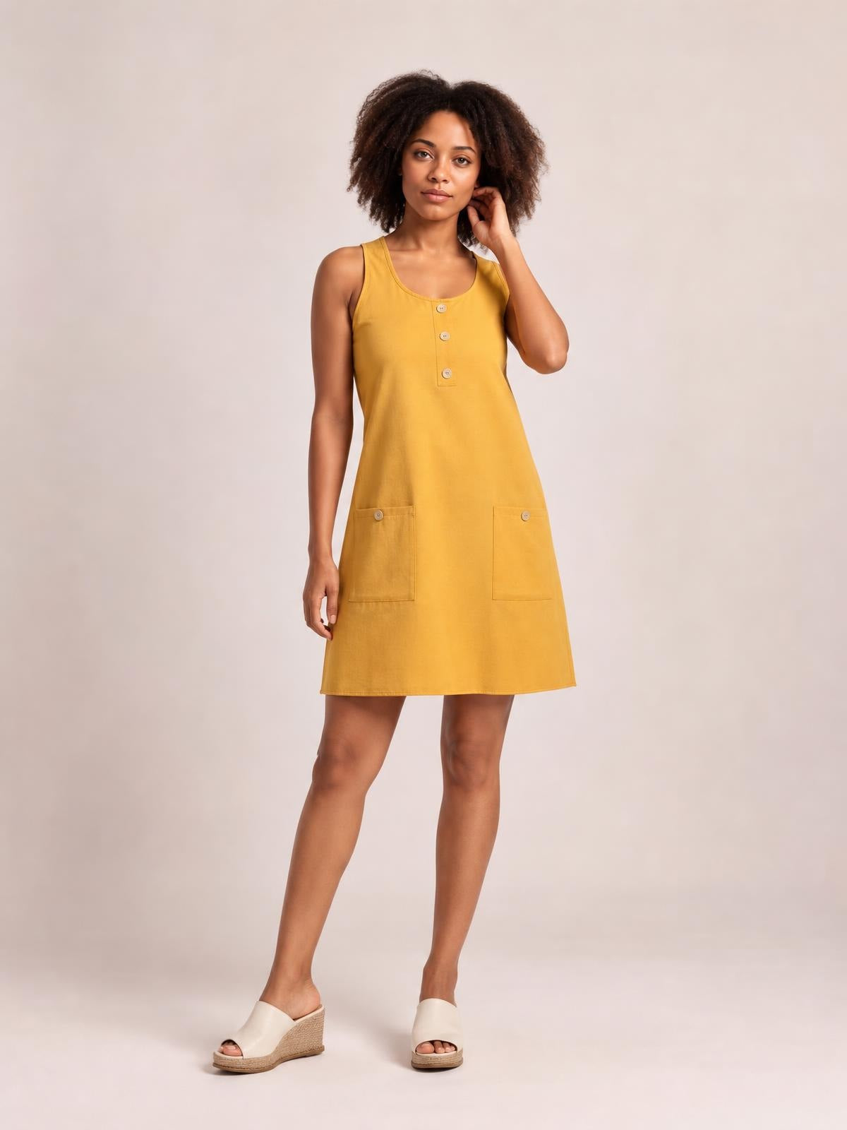A model poses in a mustard yellow round-neck mini dress with wide straps, a three-button front placket, two patch pockets, and a relaxed A-line silhouette.