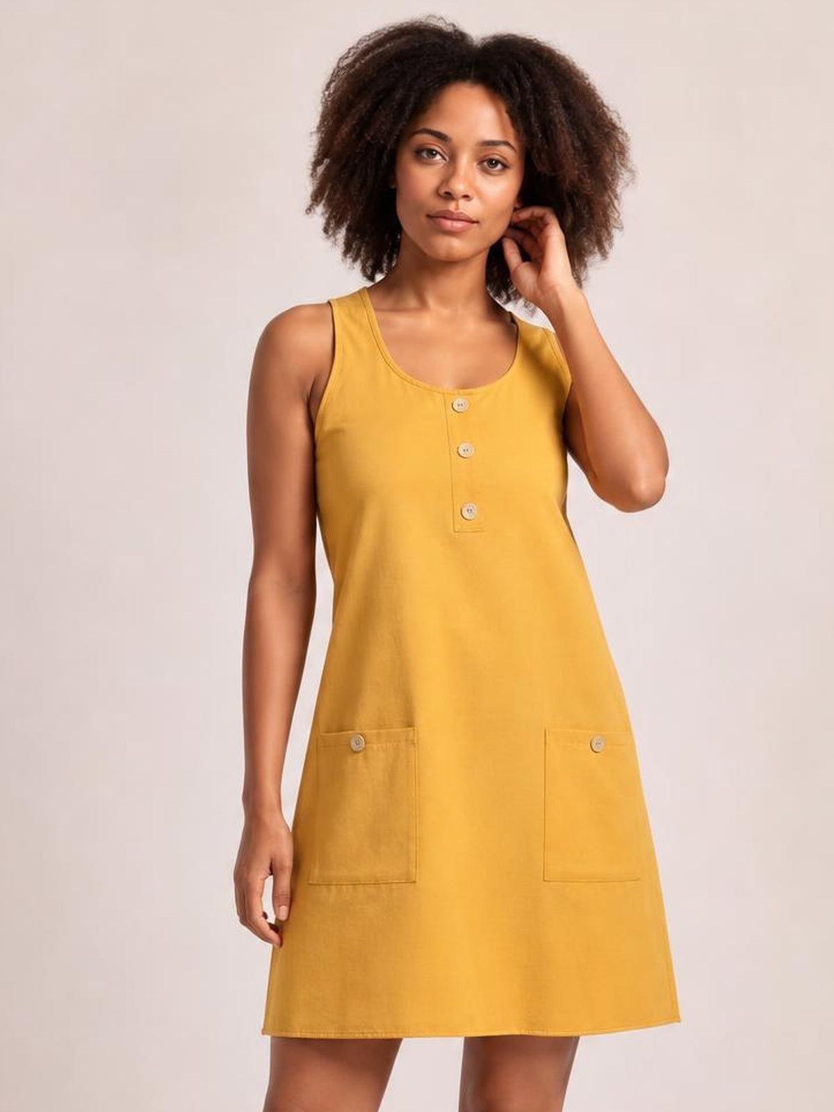Model poses in a mustard-yellow round neck mini dress with button detailing, wide shoulder straps, a relaxed A-line silhouette, and two front patch pockets.
