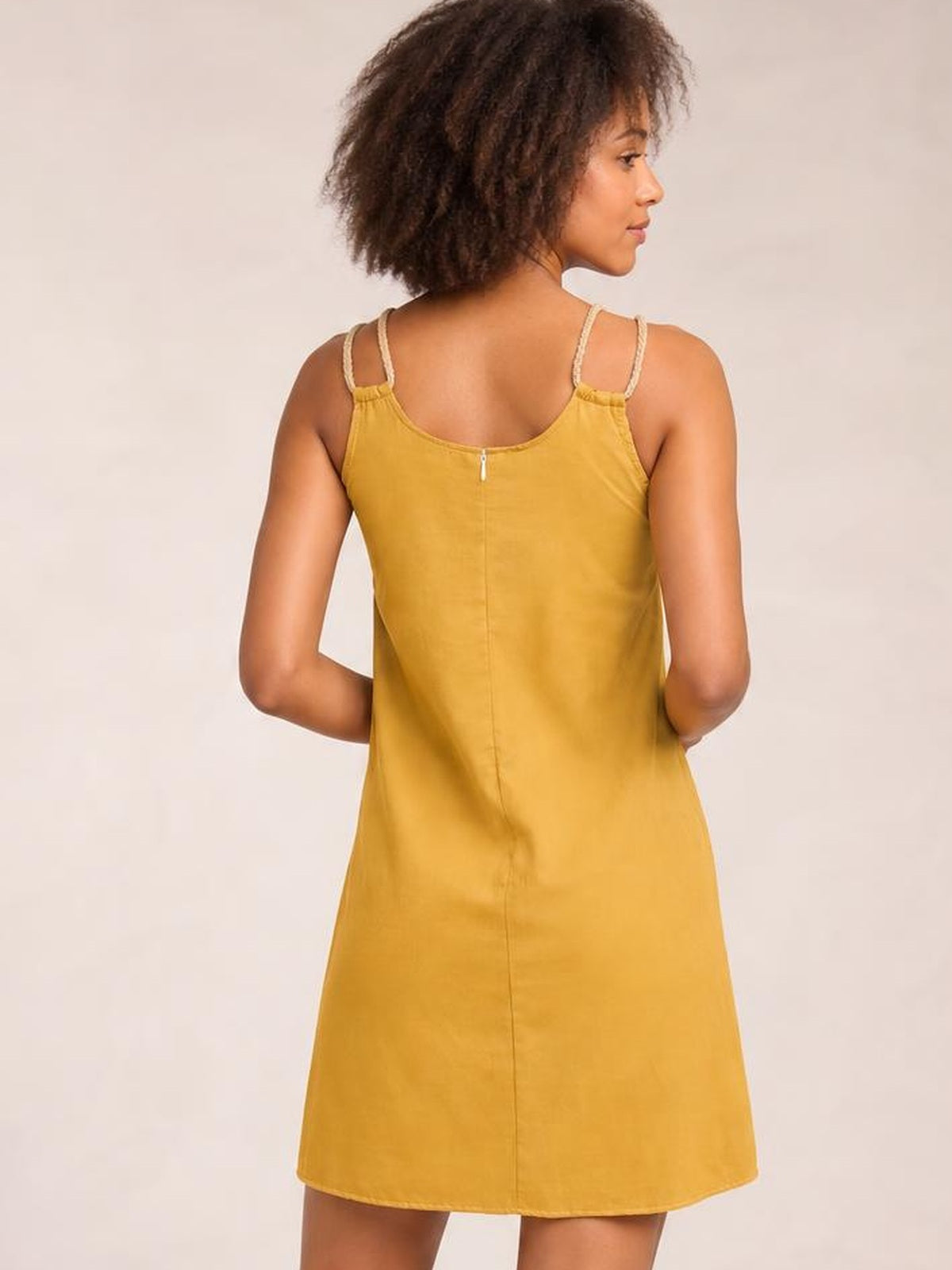 Model shown from behind in a mustard yellow mini dress with double spaghetti straps, a rounded neckline, relaxed A-line silhouette, and center-back seam.