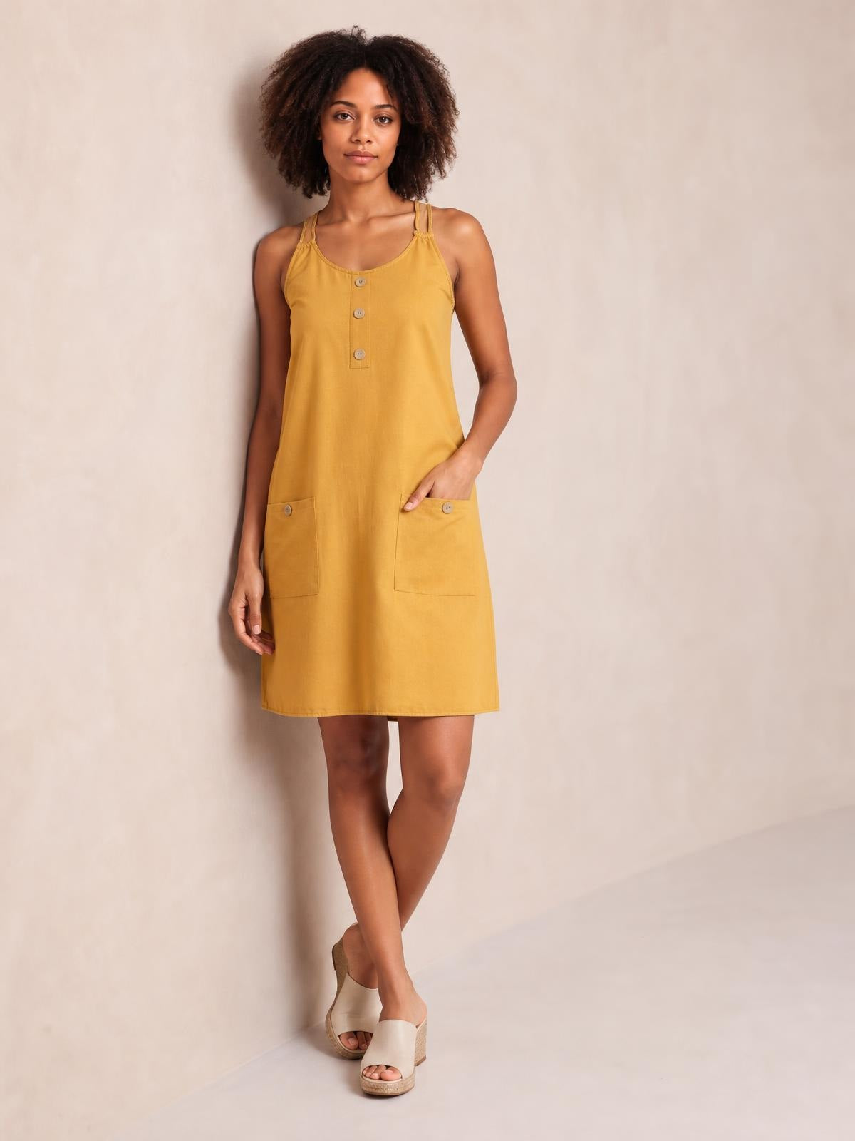 Model poses in a mustard yellow mini dress with a round neck, double spaghetti straps, front buttons, and two patch pockets.