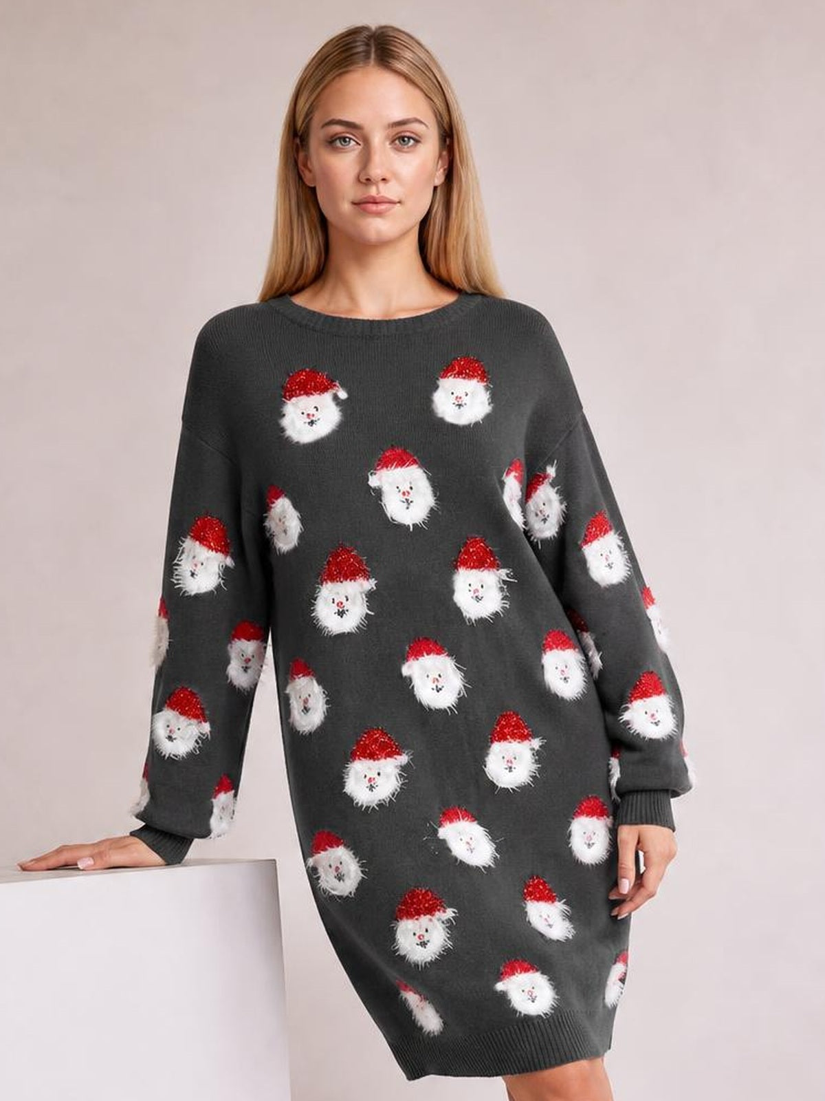 A model leans beside a pedestal wearing a dark gray round-neck sweater dress with dropped shoulders, long sleeves, ribbed cuffs and hem, and Santa face motifs.