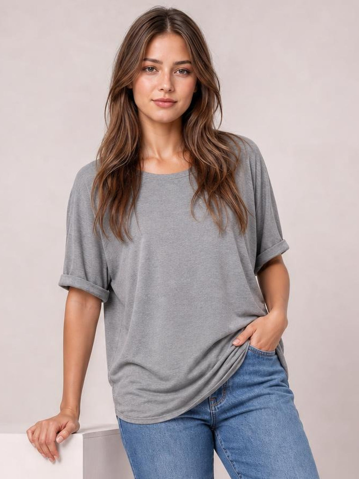 Model wearing a gray round neck half sleeve T-shirt with rolled cuffs and a relaxed silhouette, gently pulling the hem while standing in jeans.