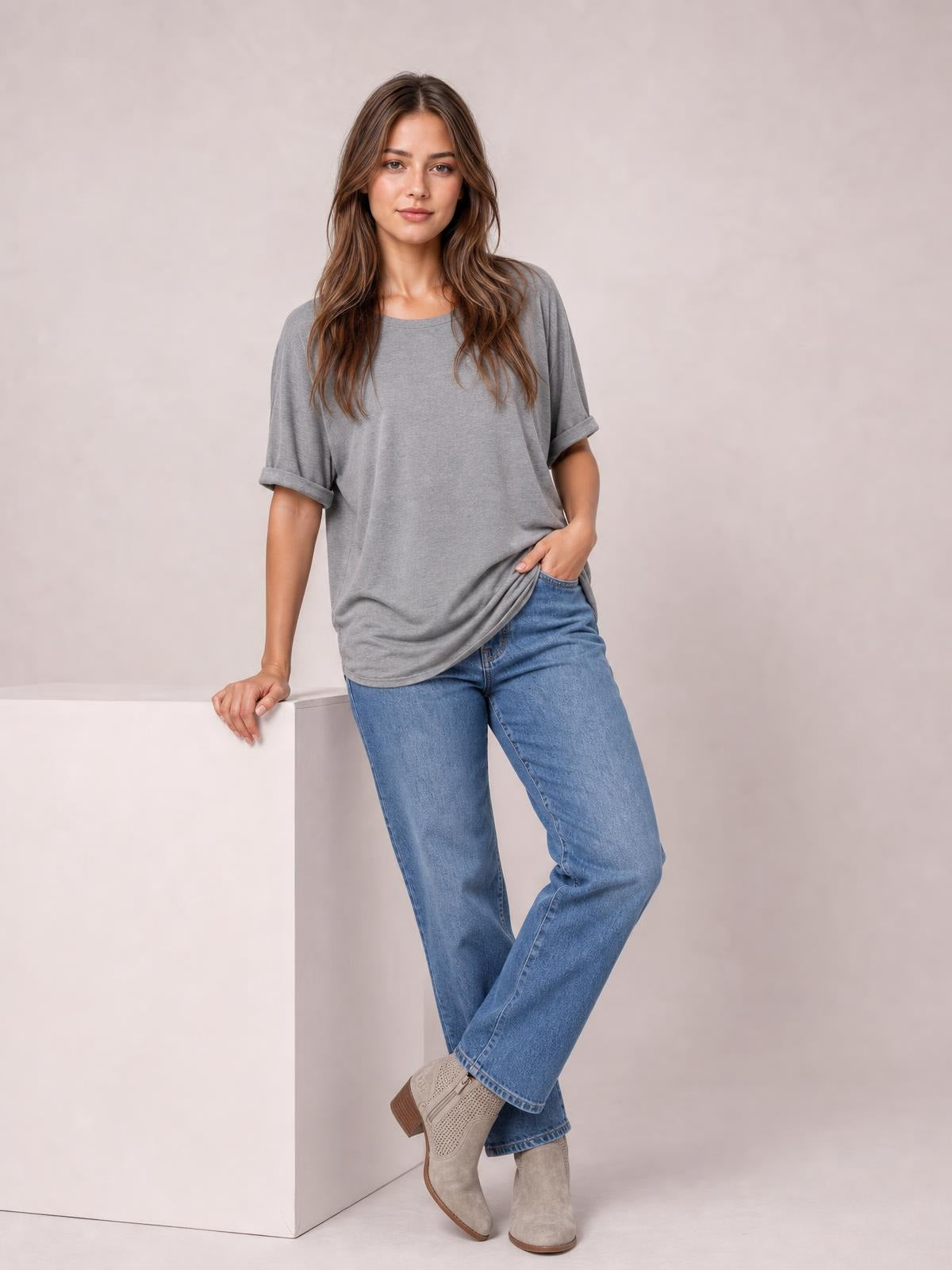 Model poses in a gray round neck half sleeve T-shirt with rolled cuffs and a relaxed, slightly loose silhouette, paired with jeans.