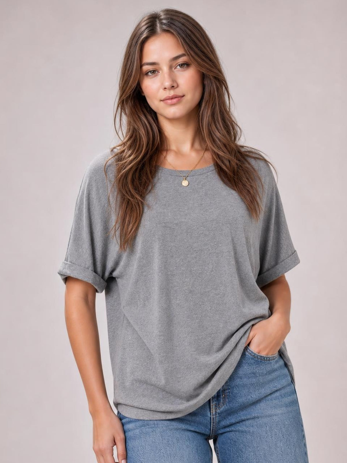 A model wears a gray round neck half sleeve T-shirt with rolled cuffs and a relaxed, slightly loose silhouette, lifting the hem over blue jeans.