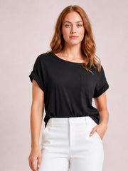 Model wearing a black round neck half sleeve T-shirt with rolled cuffs, relaxed silhouette, and visible chest pocket, posing with one hand in her pocket.