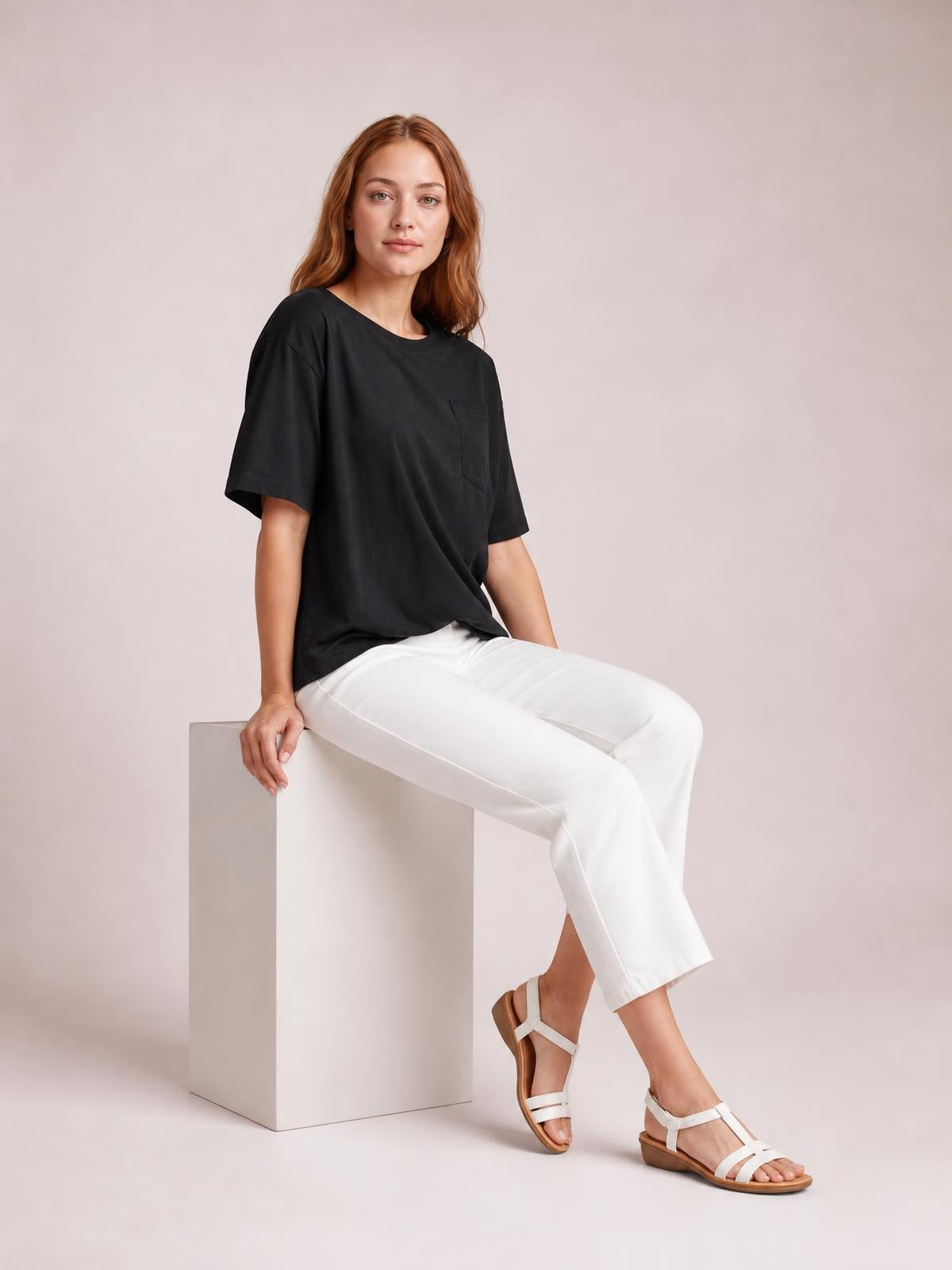 A model sits with one hand resting beside her, wearing a loose black round neck half sleeve T-shirt with a chest pocket and straight hem.