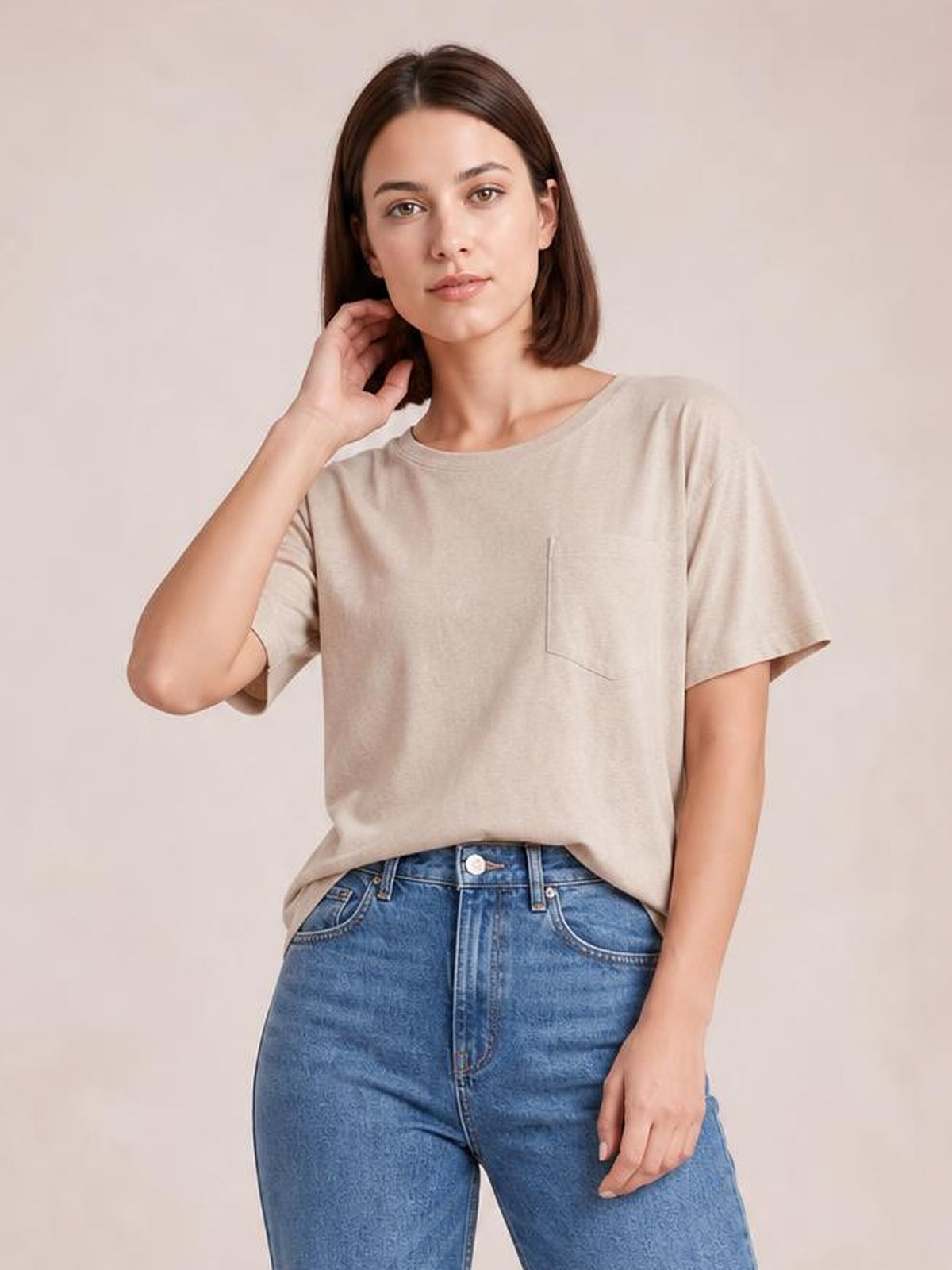 A woman poses in a beige round neck half sleeve T-shirt with a relaxed silhouette, visible chest pocket, and one hand resting near her hair.