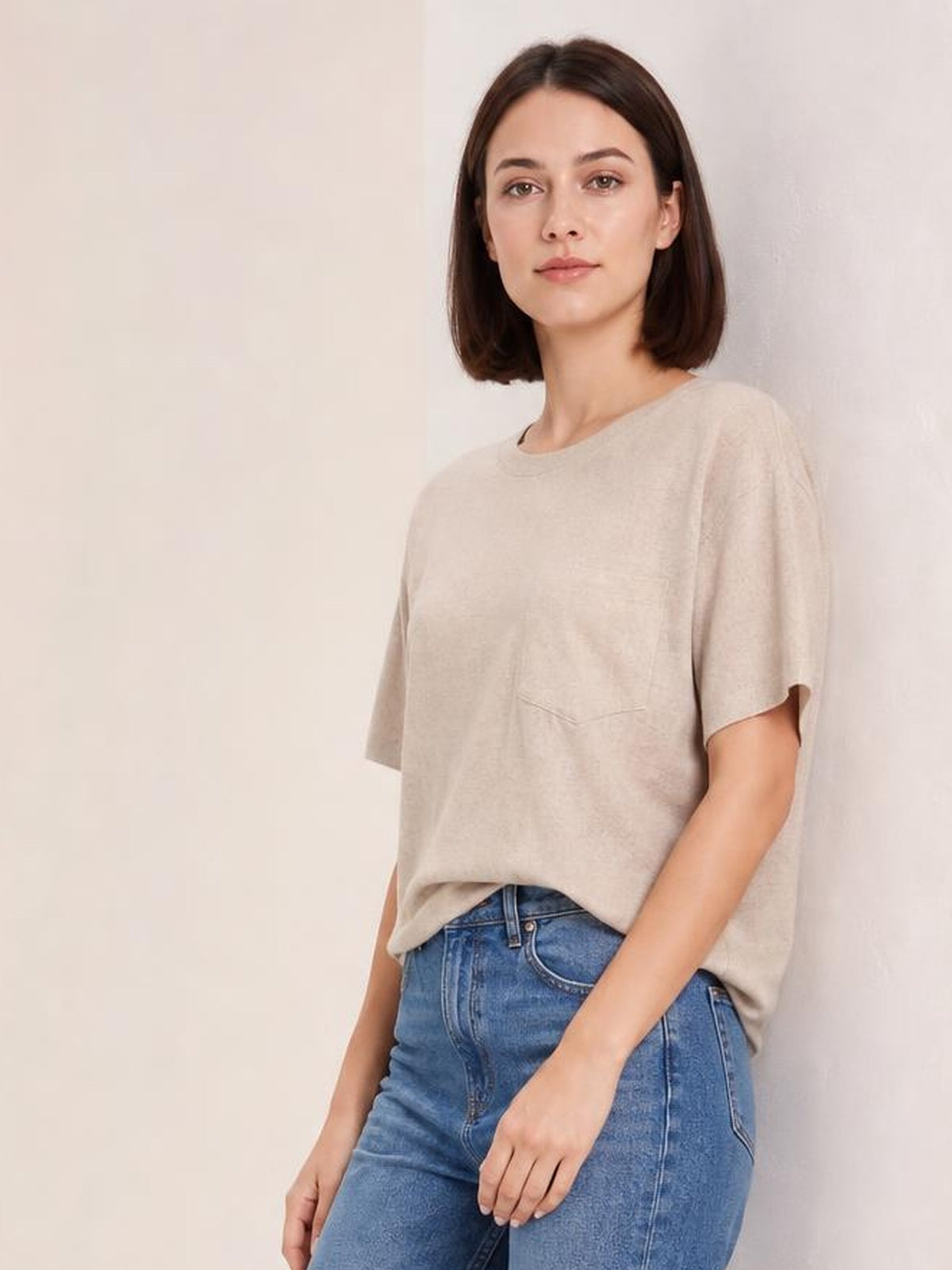 A woman wears a light beige round-neck half-sleeve T-shirt with a relaxed silhouette, subtle front pocket, and slightly dropped hem over blue jeans.