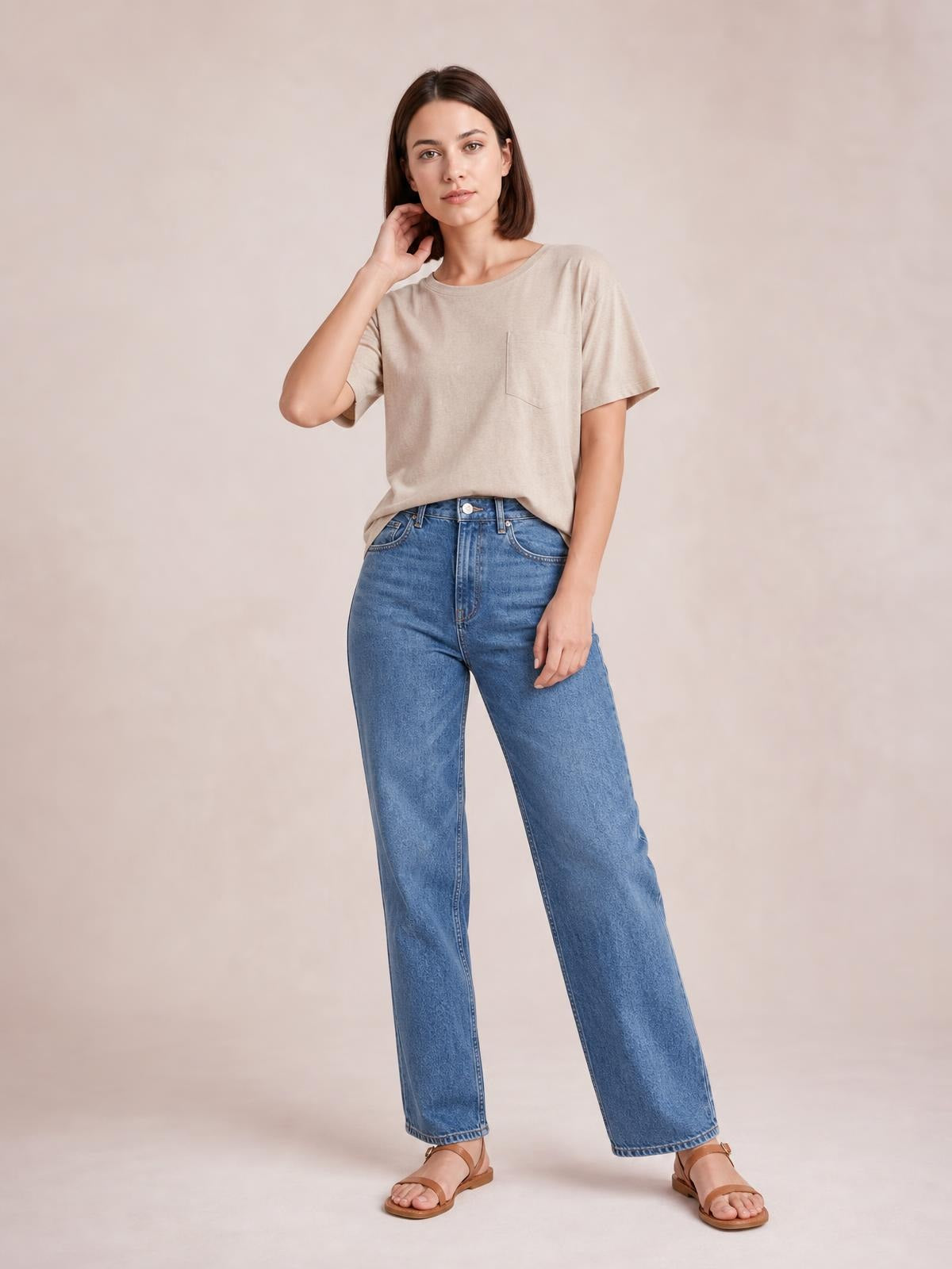 Model wears a beige round neck half sleeve T-shirt with a relaxed silhouette and chest pocket, posing with one hand near her hair and blue jeans.