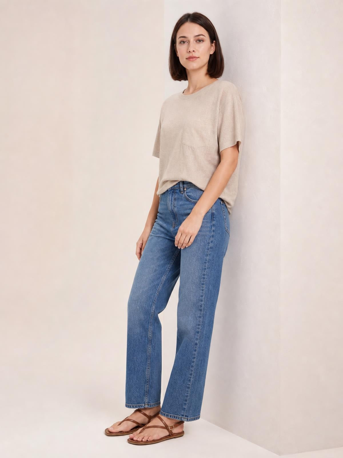 Woman wearing a beige round neck half sleeve T-shirt with a chest pocket and relaxed silhouette, posed standing in wide-leg blue jeans.