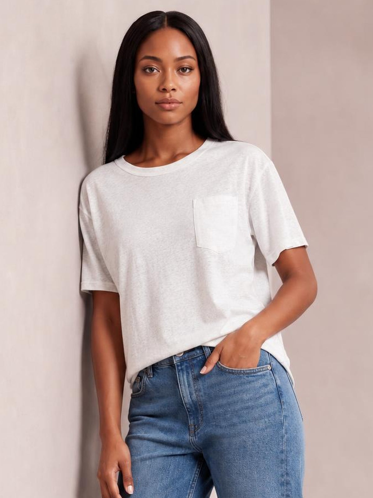 Model wears a white round neck half sleeve T-shirt with a relaxed silhouette, opaque fabric, and a visible chest pocket while posing with one hand in her jeans pocket.