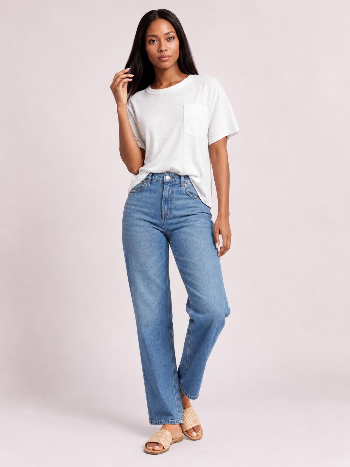 Model in a relaxed pose wears a white round neck half sleeve T-shirt with a chest pocket and loose silhouette, tucked into high-waisted blue jeans.
