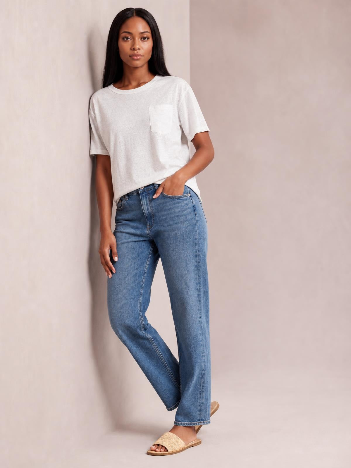 Model wearing an opaque white round neck half sleeve T-shirt with a chest pocket and relaxed silhouette, posed in blue jeans with one hand in a pocket.
