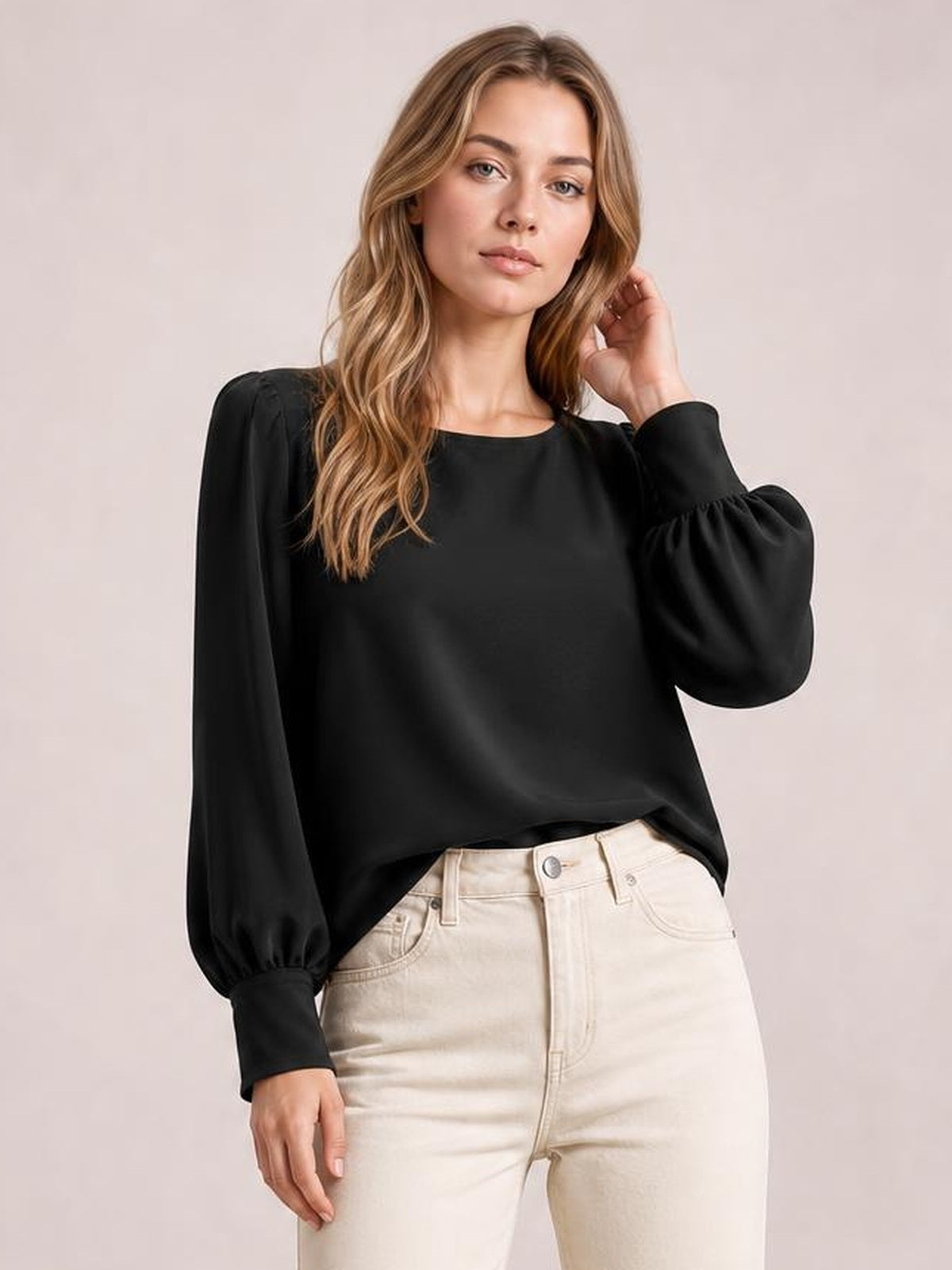 Model wearing a black round-neck blouse with voluminous lantern sleeves, gathered cuffs, and a relaxed silhouette, posing with one hand touching her hair.