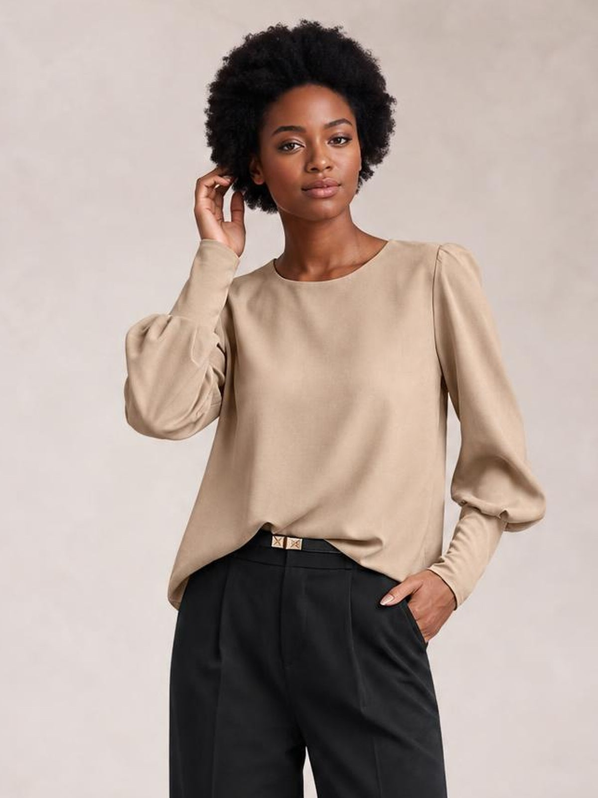 Model wearing a beige round neck blouse with voluminous lantern sleeves, a relaxed straight silhouette, and gathered cuffs, posing with one hand in her pocket.