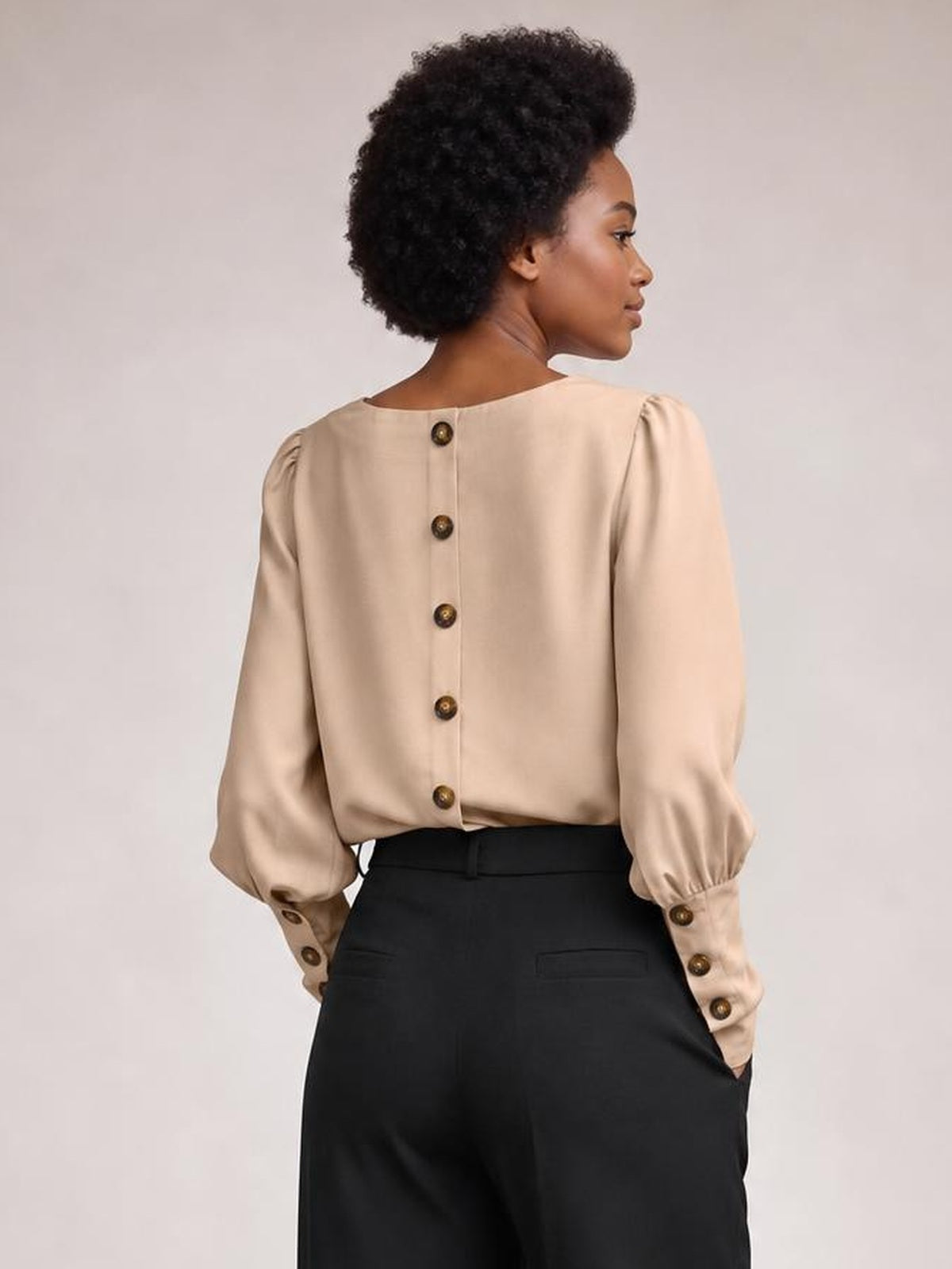 A model shows the back of a beige round-neck blouse with voluminous lantern sleeves, button-front construction, gathered cuffs, and a relaxed silhouette.