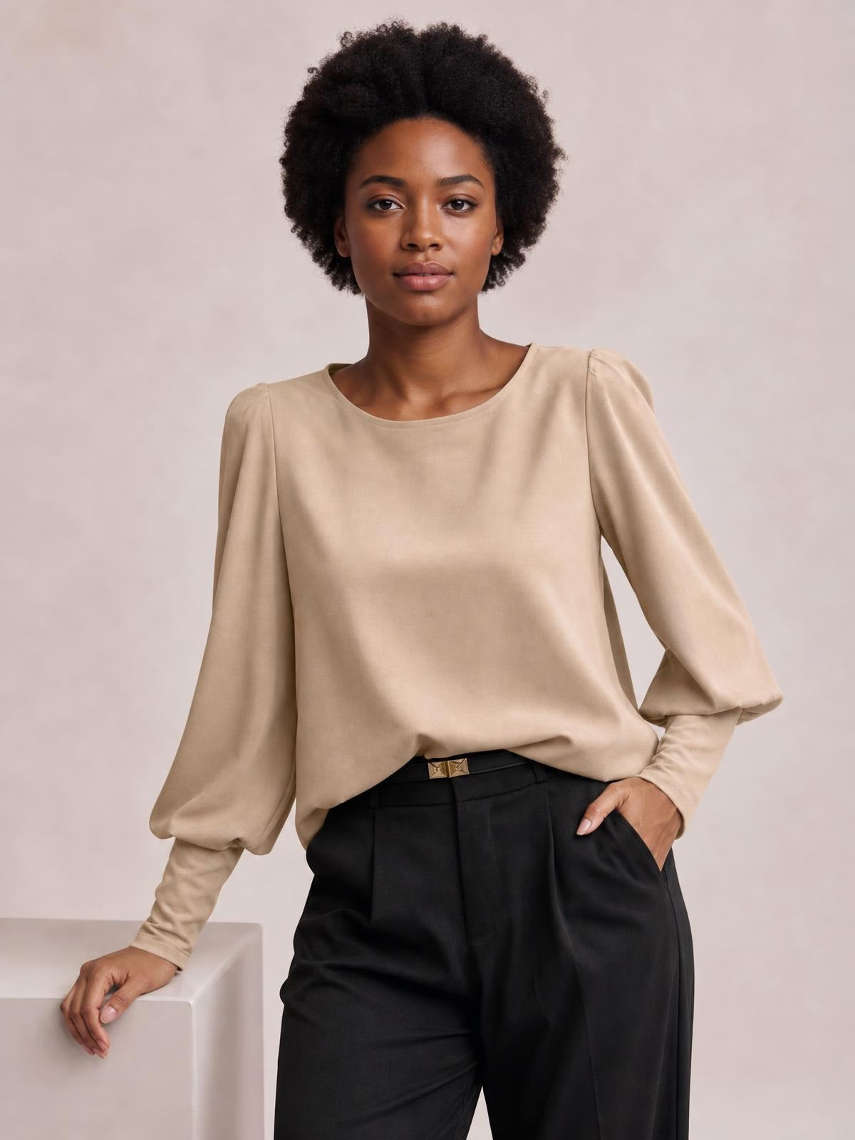 Model poses in a beige round neck blouse with voluminous lantern sleeves, gathered cuffs, and a relaxed straight silhouette, styled with black trousers.