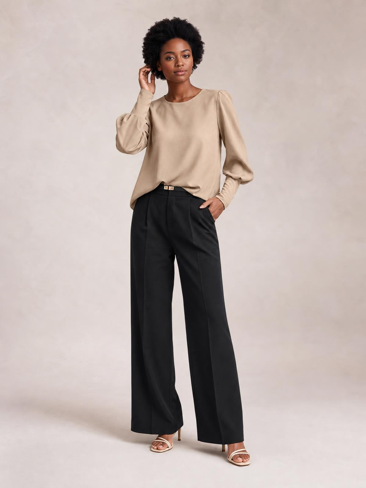 Model wearing a beige round neck blouse with gathered lantern sleeves, a relaxed straight silhouette, and long cuffs, posed with one hand at her hair.