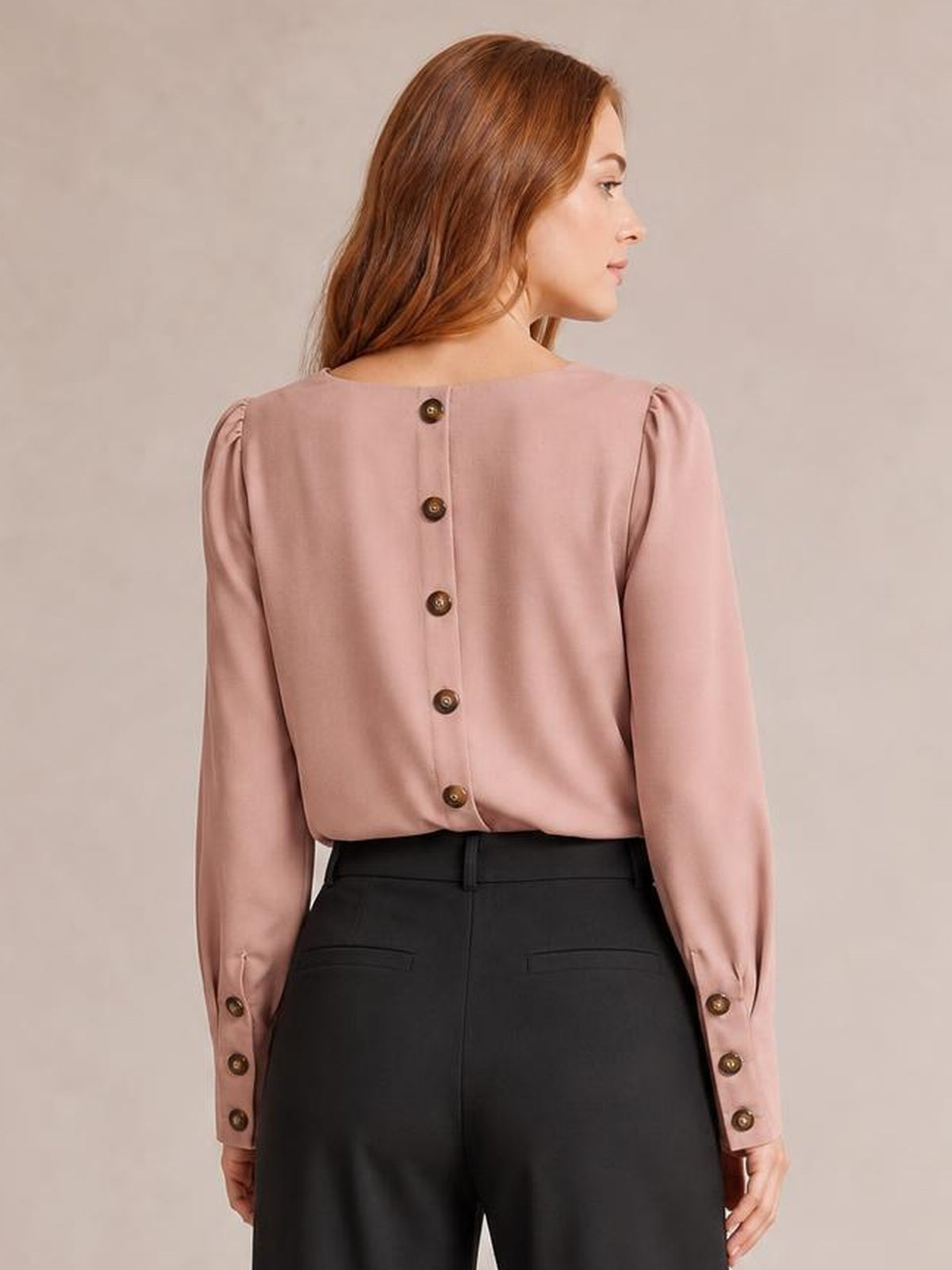 Model viewed from behind wears a dusty pink round neck blouse with lantern sleeves, button-back construction, and buttoned cuffs tucked into black trousers.