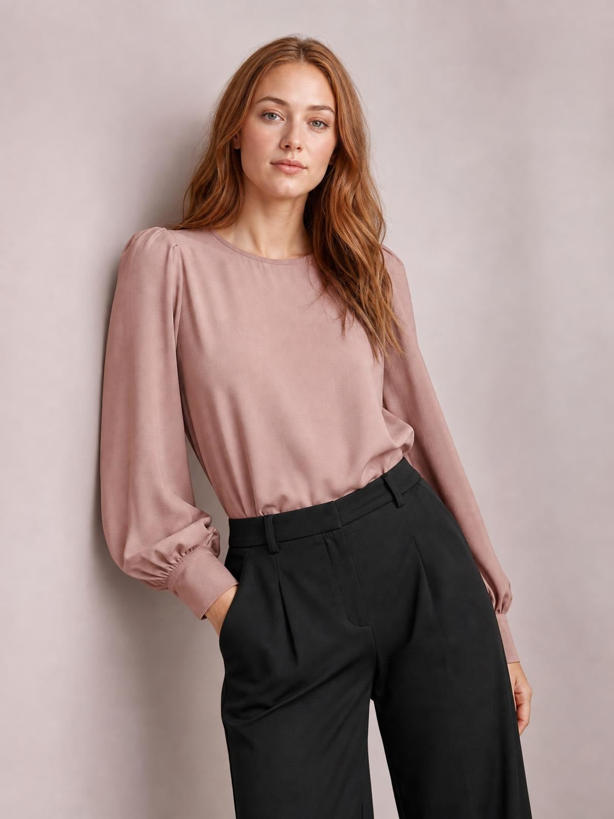 A model wears a dusty pink round-neck lantern sleeve blouse with a relaxed silhouette and gathered cuffs, tucked into black trousers as one hand rests in a pocket.