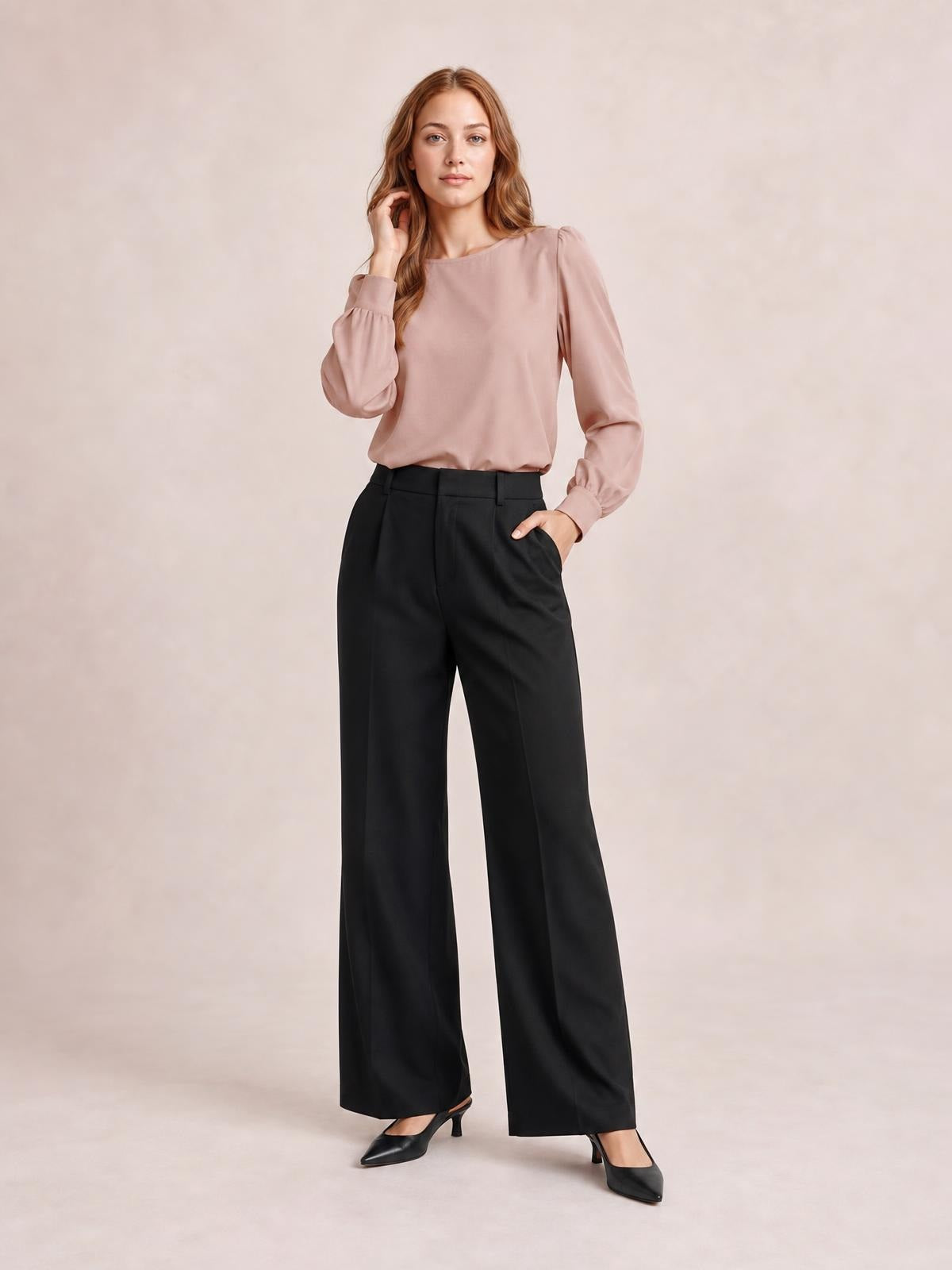 Model poses in a blush pink round-neck blouse with voluminous lantern sleeves, gathered cuffs, and a softly relaxed silhouette tucked into black wide-leg trousers.