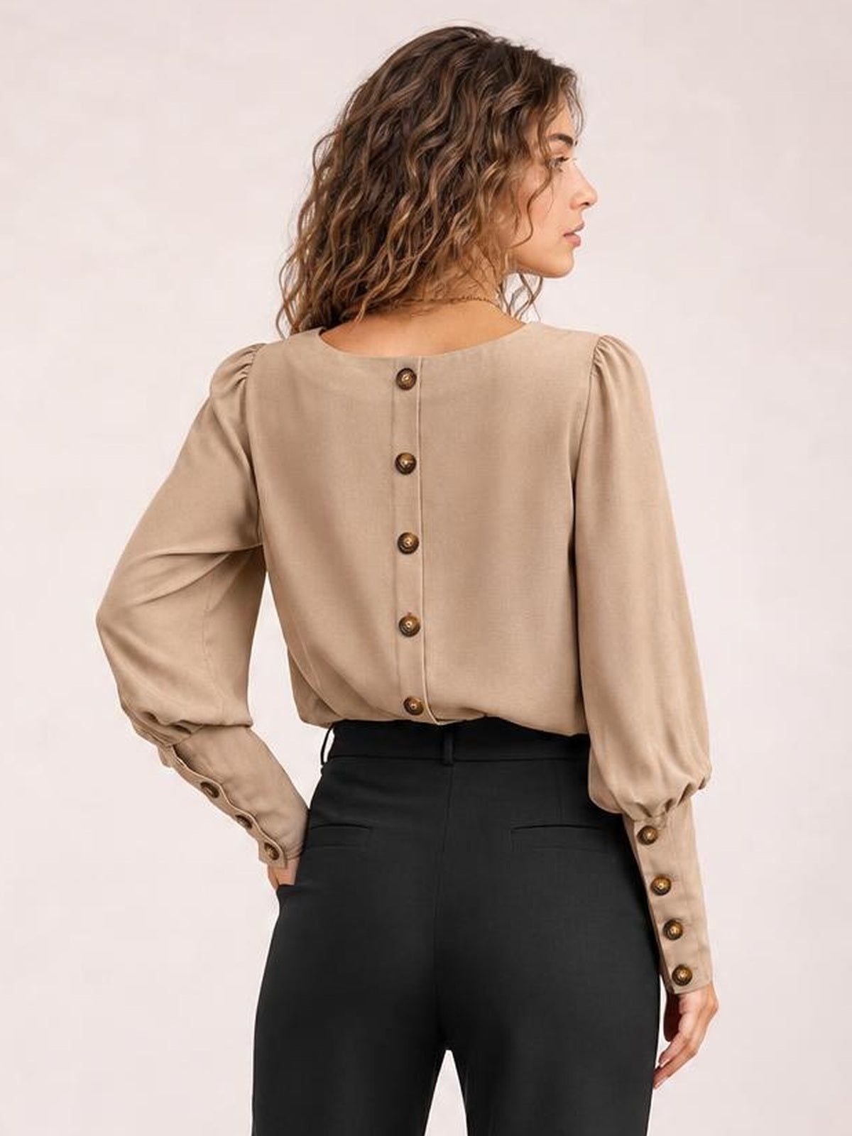 Model shown from the back wearing a beige round-neck blouse with voluminous lantern sleeves, buttoned back closure, and buttoned cuffs, hands at her hips.