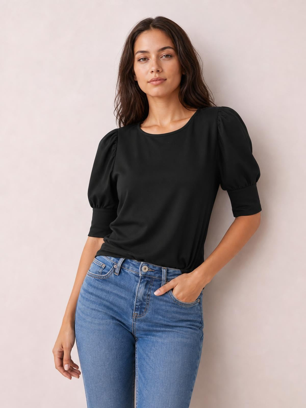 A model wears a black round-neck lantern sleeve blouse with gathered shoulders, voluminous sleeves, fitted cuffs, and a relaxed silhouette while posing with one hand in her jeans pocket.