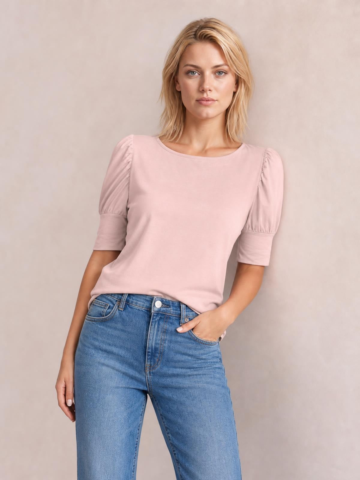 Model wearing a pink round neck lantern sleeve blouse with gathered puff sleeves, fitted cuffs, and a relaxed silhouette, posed with one hand in her jeans pocket.