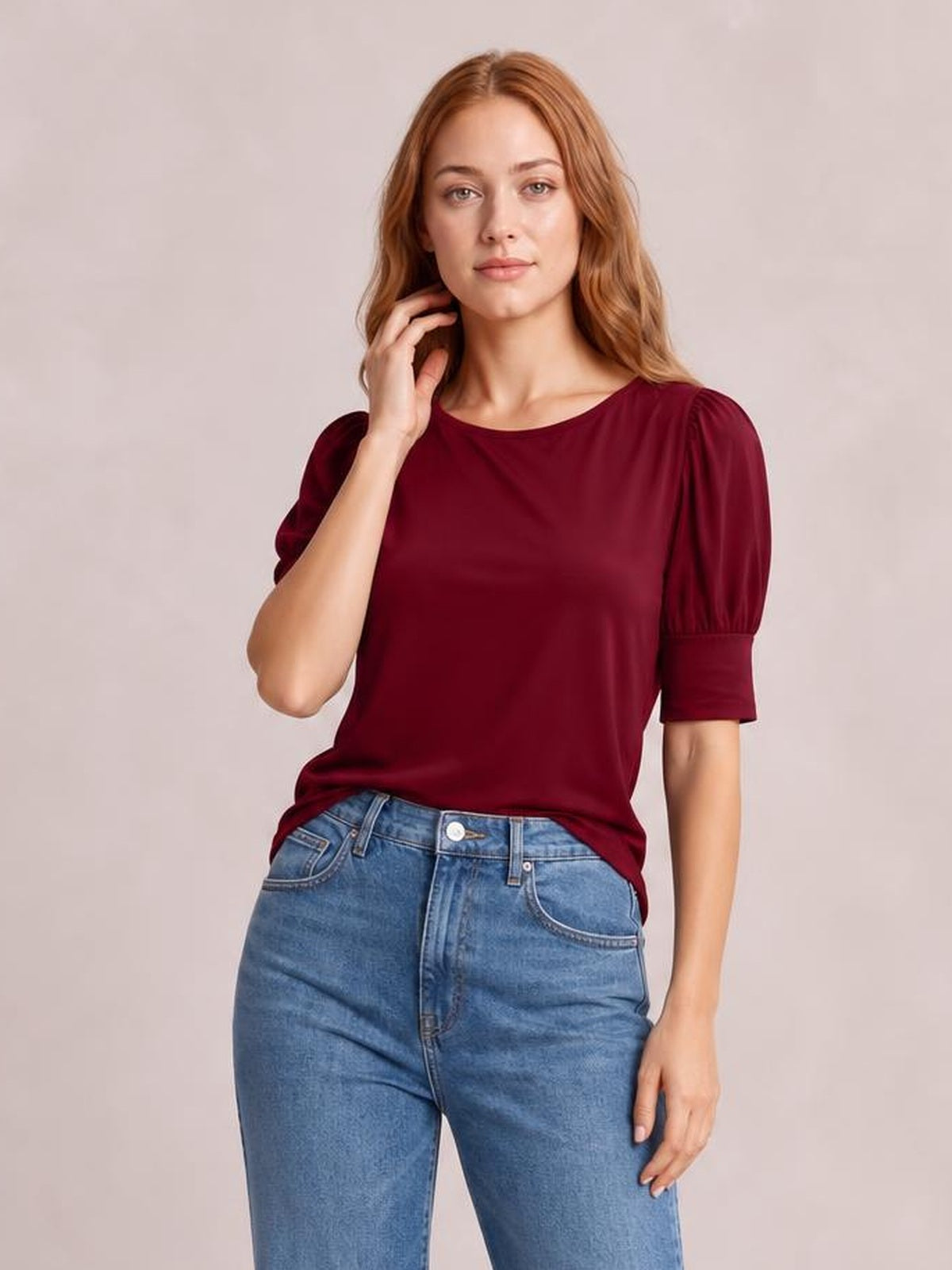 Model wearing a burgundy round neck blouse with gathered lantern sleeves and a relaxed silhouette, posing with one hand near her hair and paired with jeans.
