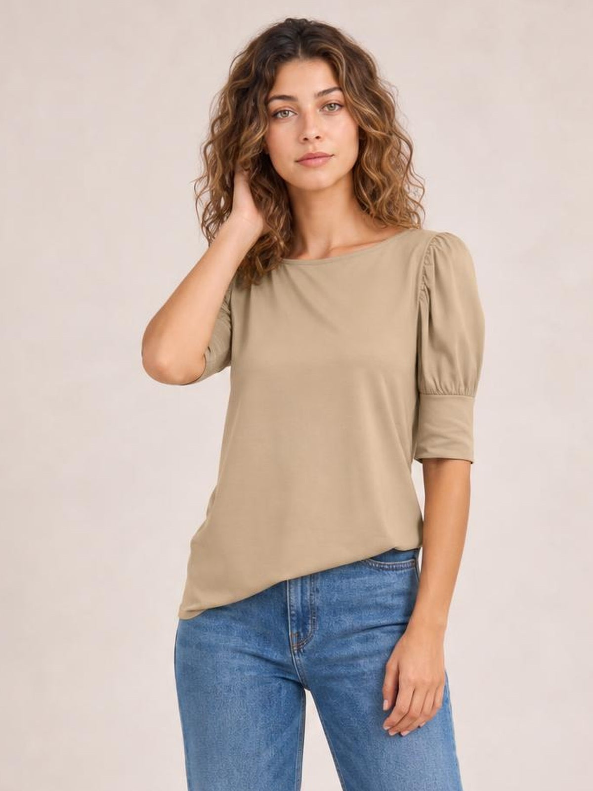 Model poses in a taupe round neck blouse with gathered lantern sleeves, a relaxed straight silhouette, and curved hem tucked into blue jeans.