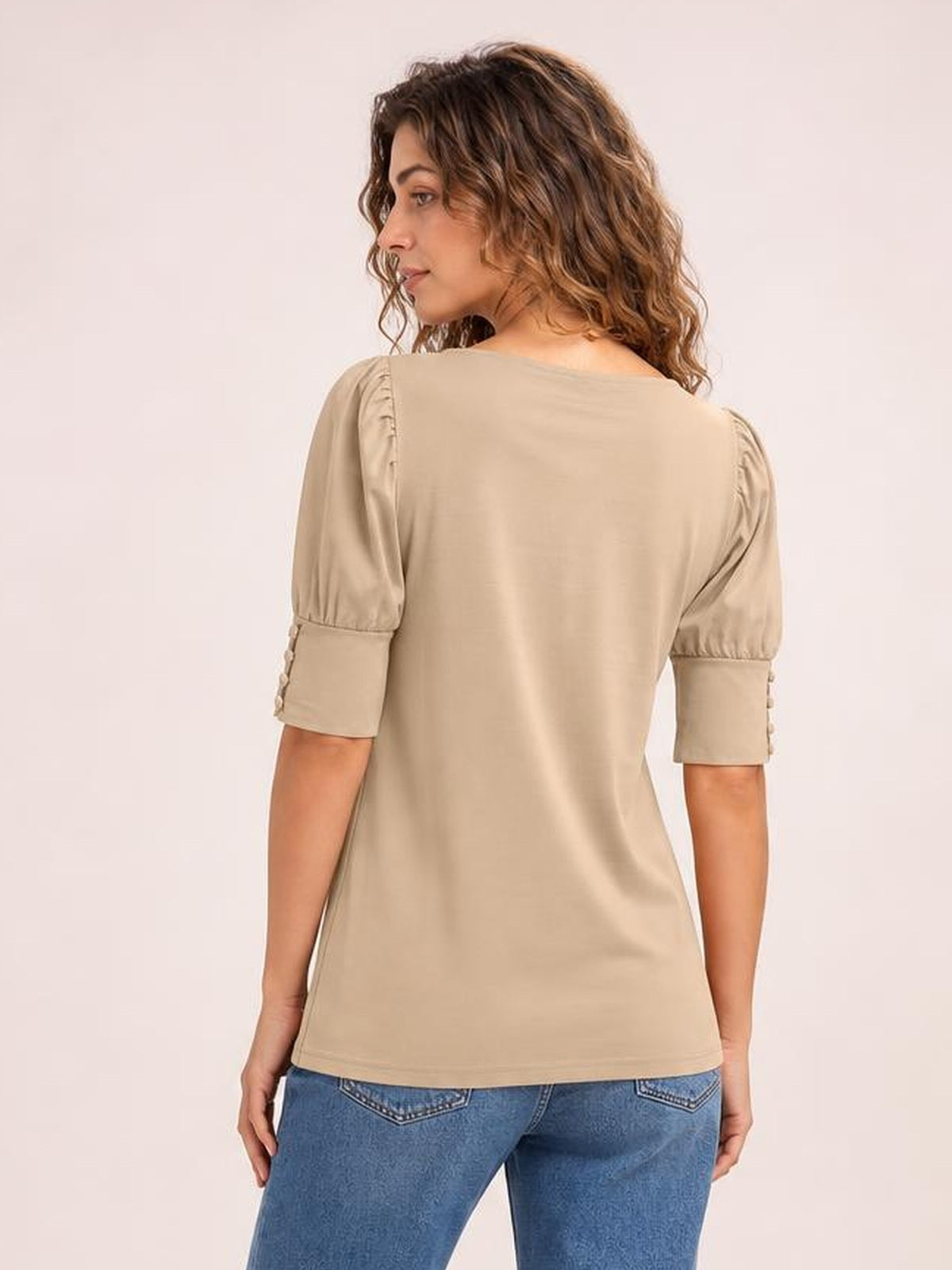 A model looks over her shoulder in a beige round-neck blouse with lantern sleeves, gathered puffed shoulders, cuff details, and a relaxed straight silhouette.