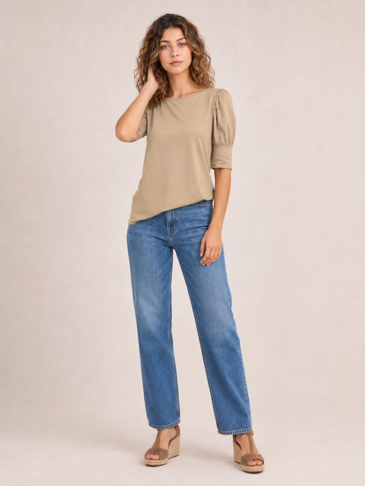 Model wearing a taupe round-neck blouse with gathered lantern sleeves and a relaxed straight silhouette, posing with one hand in her hair.
