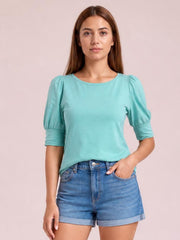 A front-facing model wears an aqua round-neck blouse with gathered lantern sleeves, a straight silhouette, and fitted three-quarter cuffs, resting one hand in her shorts pocket.