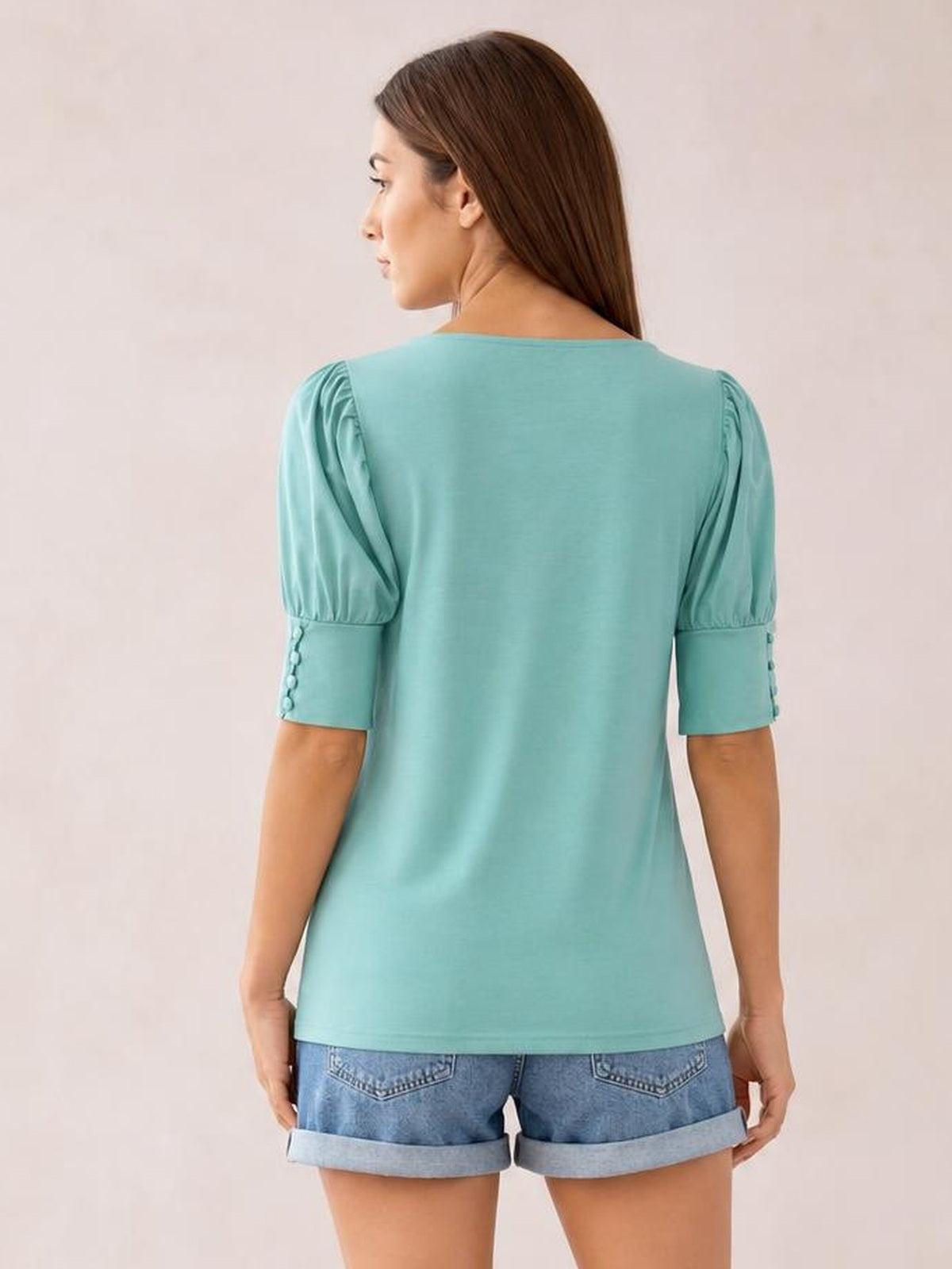 A model stands with her back to the camera wearing a turquoise round-neck blouse with gathered lantern sleeves, cuffed button details, and a relaxed straight silhouette.