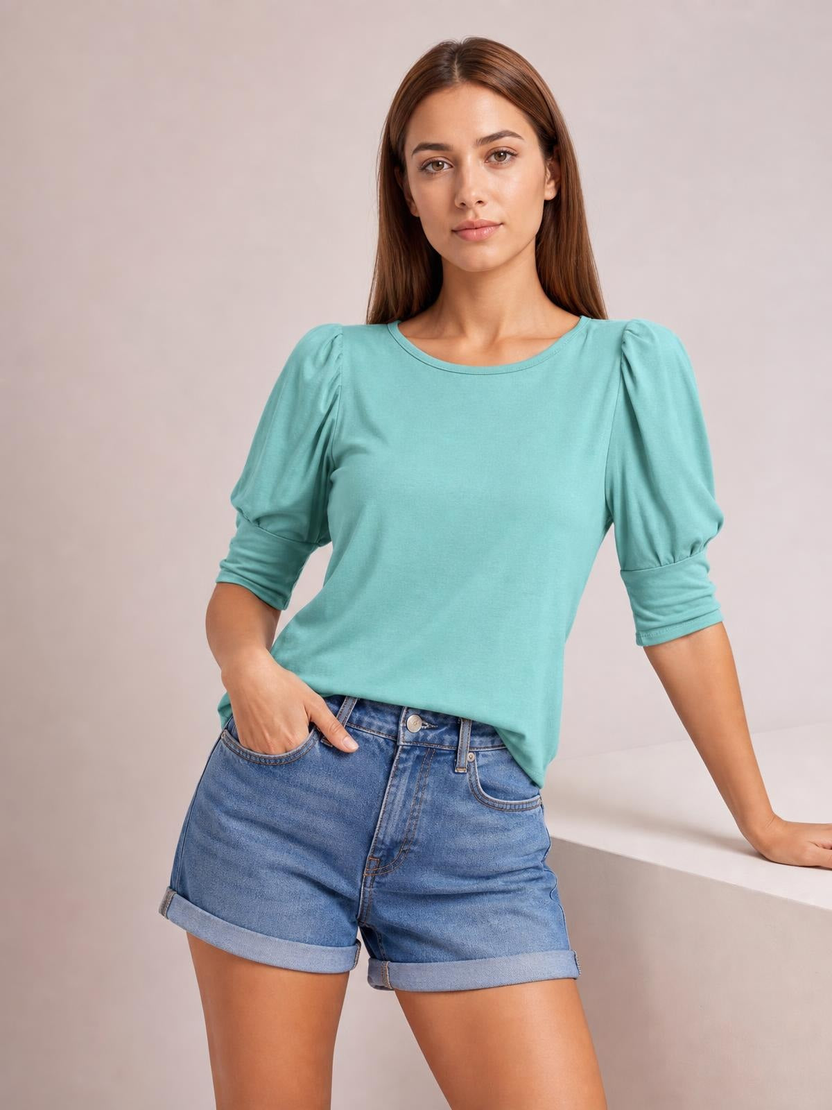 Model wears a turquoise round neck lantern sleeve blouse with a relaxed silhouette, gathered shoulders, and ruched three-quarter sleeves while posing with one hand in her shorts pocket.