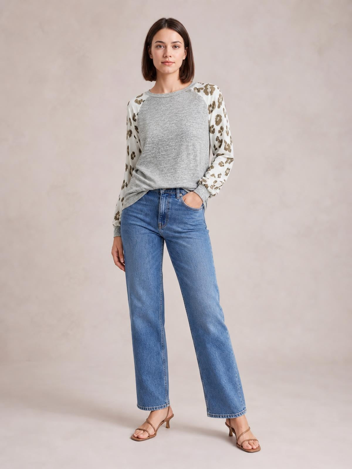 A woman stands in a gray round-neck top with leopard-print raglan sleeves, a relaxed silhouette, ribbed cuffs, and one hand tucked into her jeans pocket.