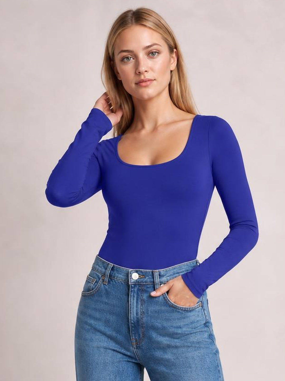 Model wearing a royal blue round neck long sleeve bodysuit with a smooth, fitted silhouette, posing with one hand in her jeans pocket.