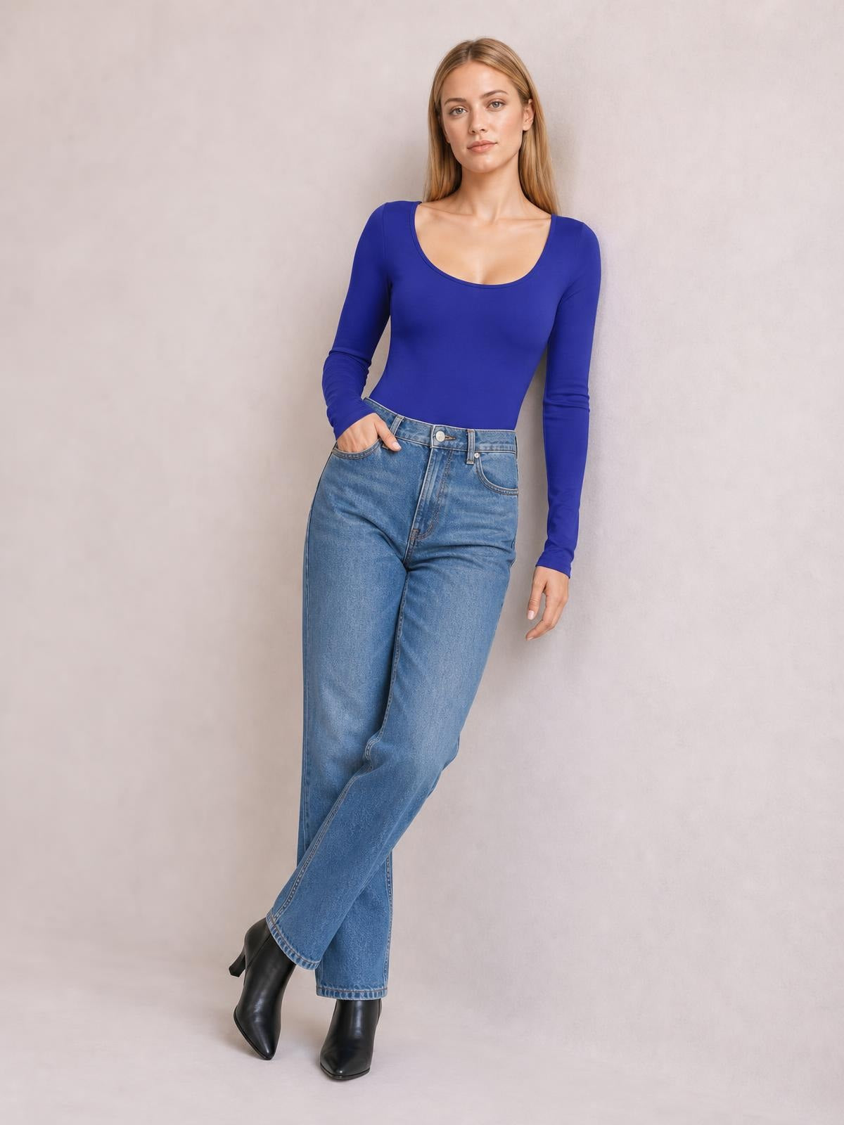 A model poses in a fitted royal-blue round-neck long-sleeve bodysuit with a smooth opaque silhouette, one hand in her jeans pocket and legs crossed.