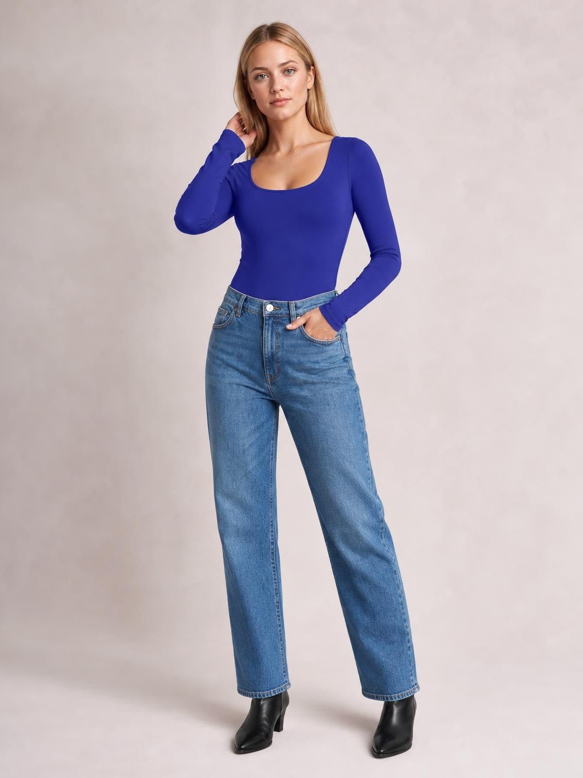 Model wears a cobalt blue round-neck long-sleeve bodysuit with a fitted silhouette and moderate stretch, posing with one hand near her hair and one in her jeans pocket.