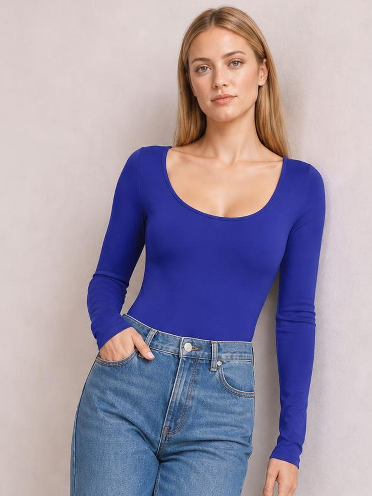 Model wearing a royal blue round neck long sleeve bodysuit with a fitted silhouette, shown tucked into jeans with one hand in a pocket.