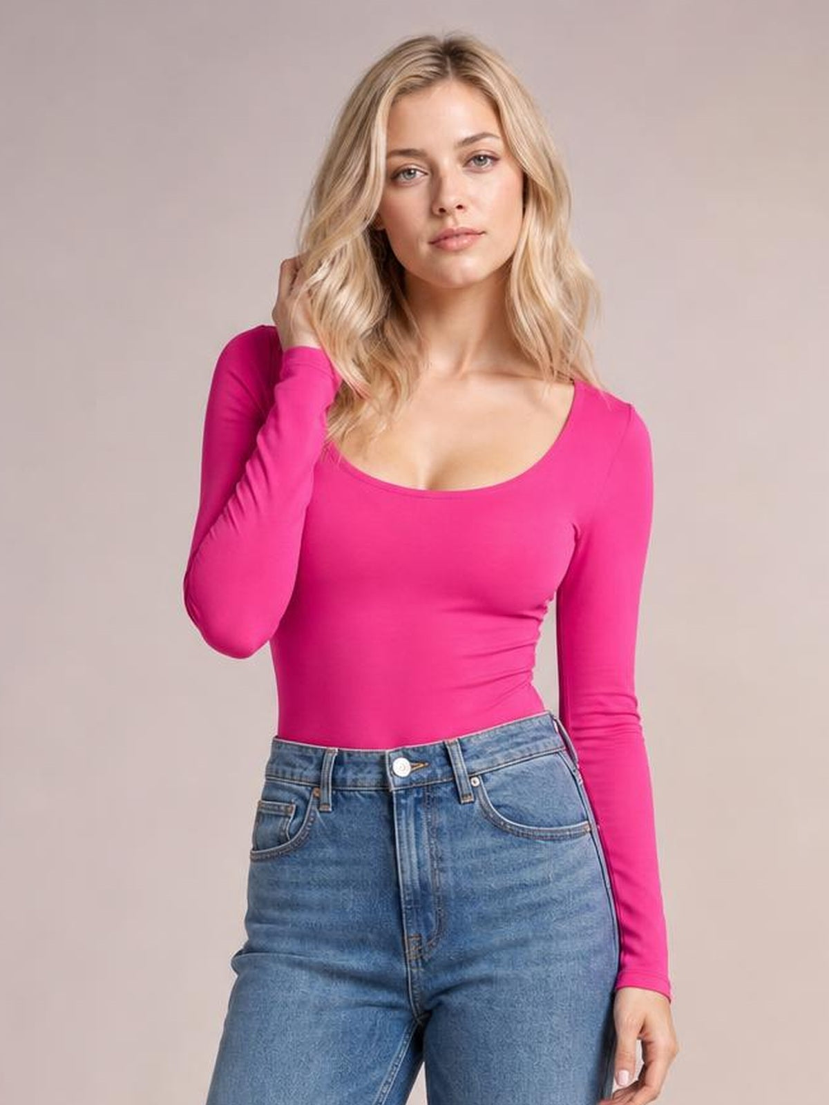 Model poses in a bright pink round neck long sleeve bodysuit with a fitted silhouette and moderate stretch, paired with blue jeans.