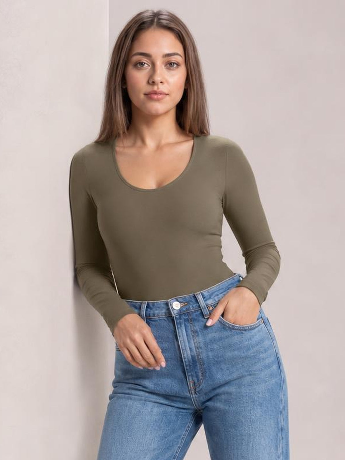 Model wears an olive round neck long sleeve bodysuit with a fitted silhouette, smooth construction, and moderate stretch, paired with jeans.