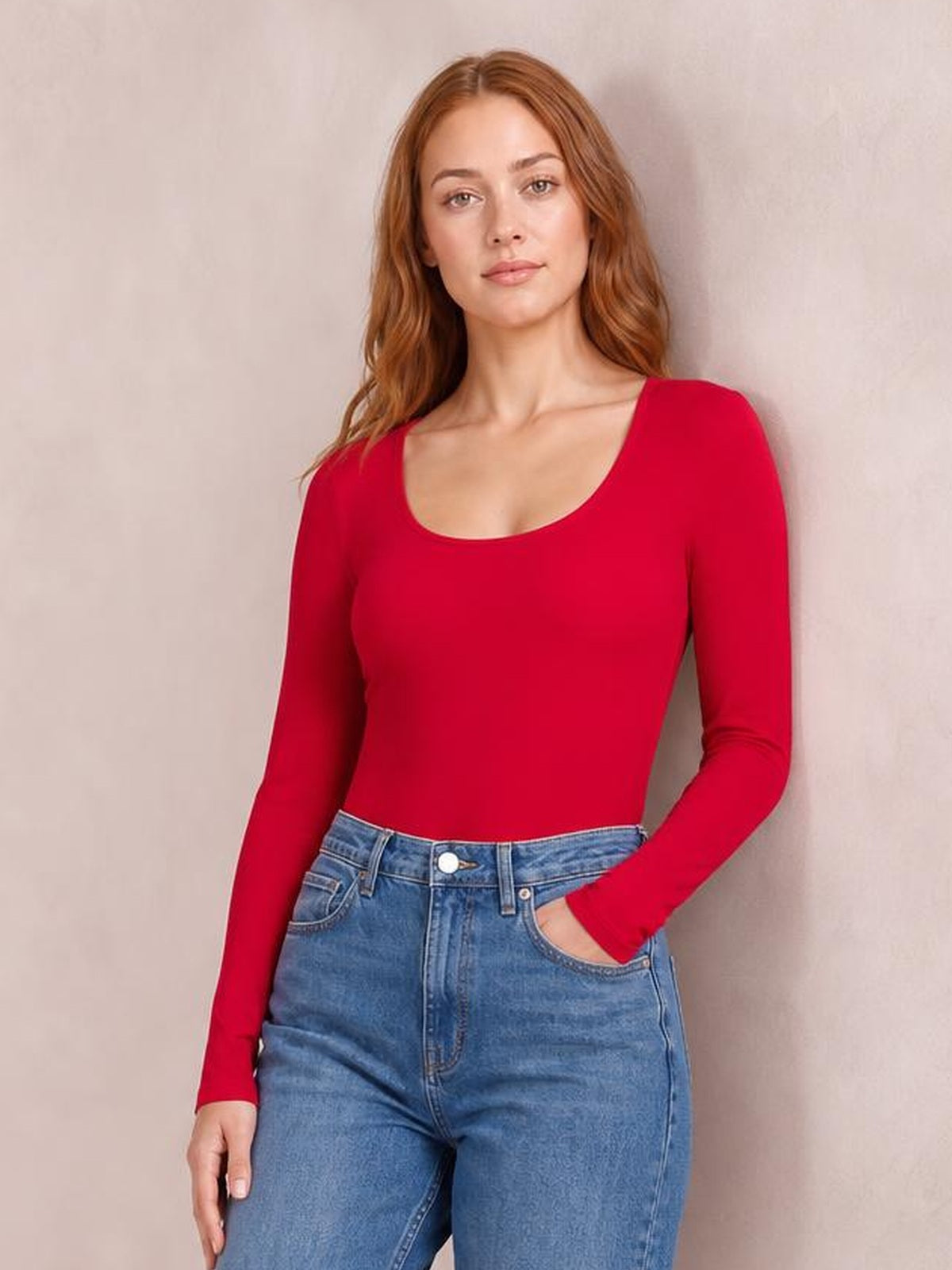 A model poses in a fitted red round neck long sleeve bodysuit with a smooth, opaque silhouette tucked into blue jeans.