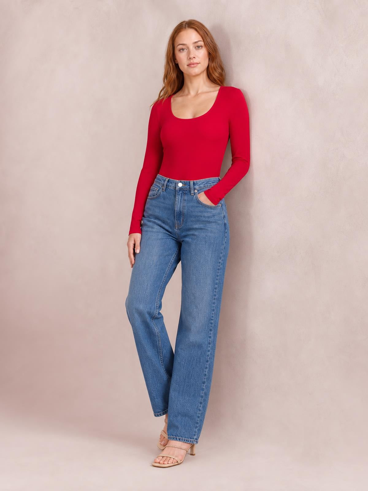 Model wearing a red round neck long sleeve bodysuit with a fitted silhouette and moderate stretch, paired with blue jeans, one hand in a pocket.