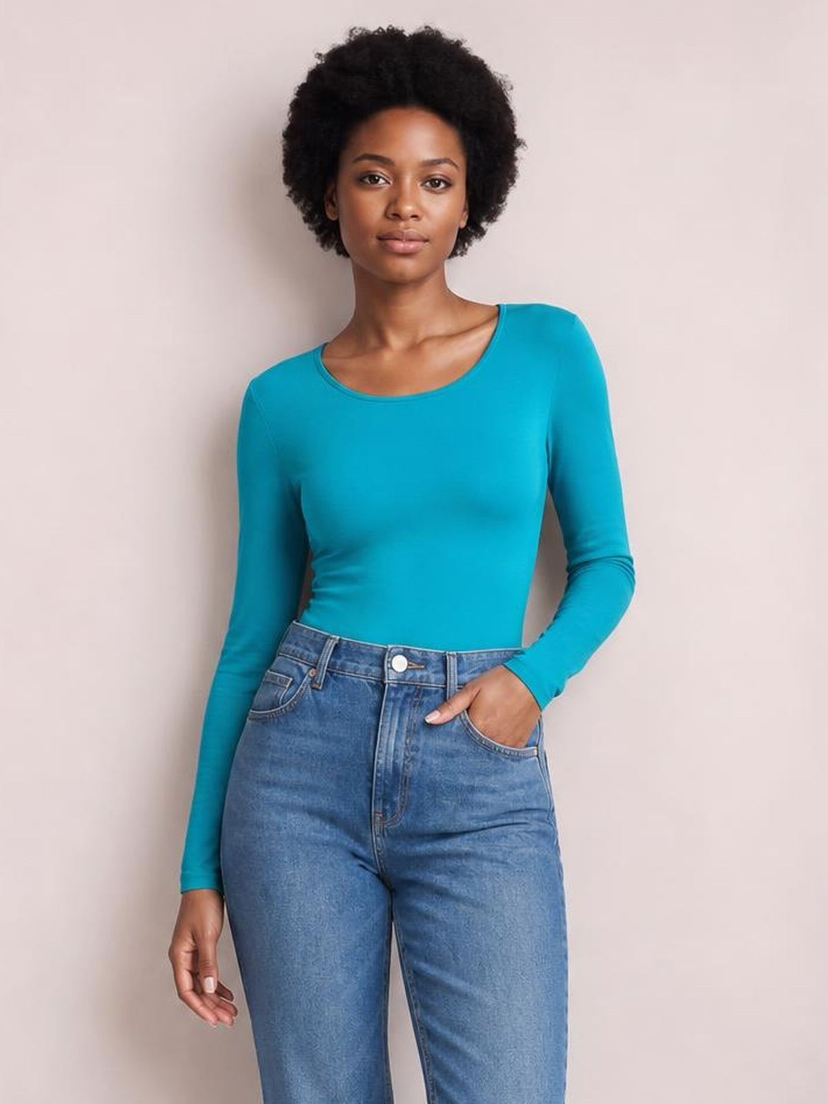 Model poses in a turquoise round neck long sleeve bodysuit with a smooth, fitted silhouette, tucked into high-waisted blue jeans.