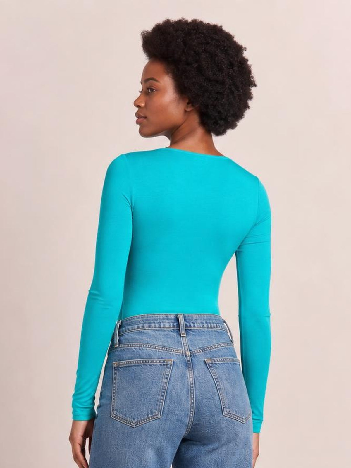 Model in a back-facing pose wears a bright turquoise round neck long sleeve bodysuit with a smooth, fitted silhouette and moderate stretch.