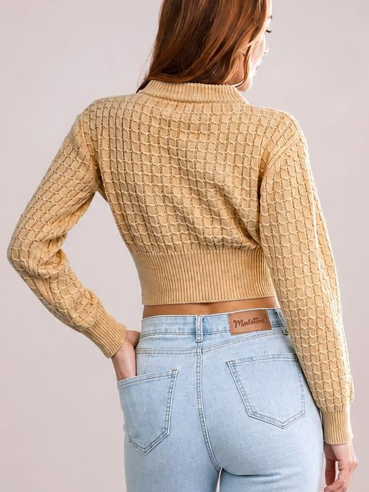 Model seen from behind wearing a tan cropped sweater with long sleeves, textured knit construction, ribbed collar, cuffs, and hem, posing with one hand in a pocket.